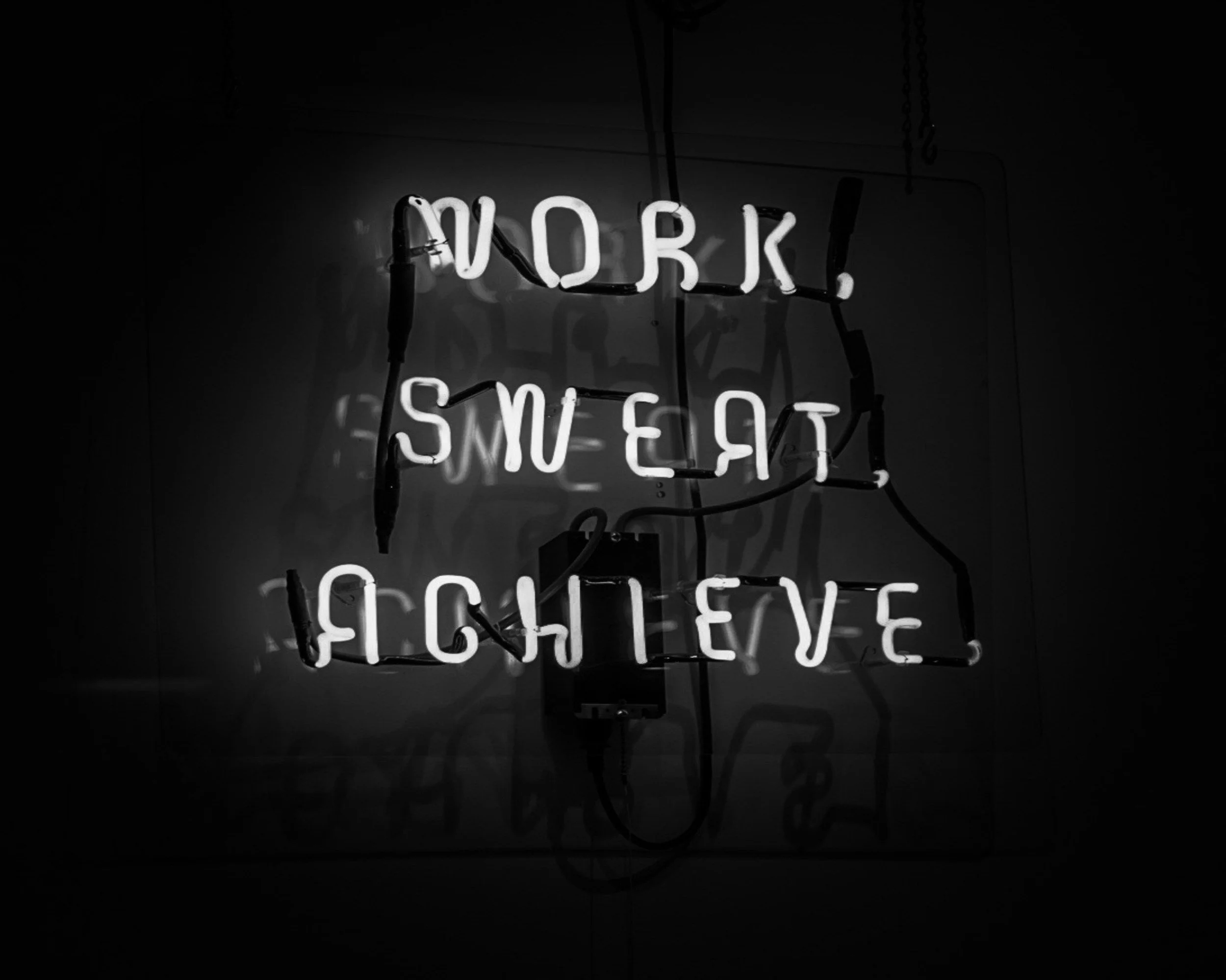 Neon sign with the quote, "Work, Sweat, Achieve" in white light, set against a dark background.