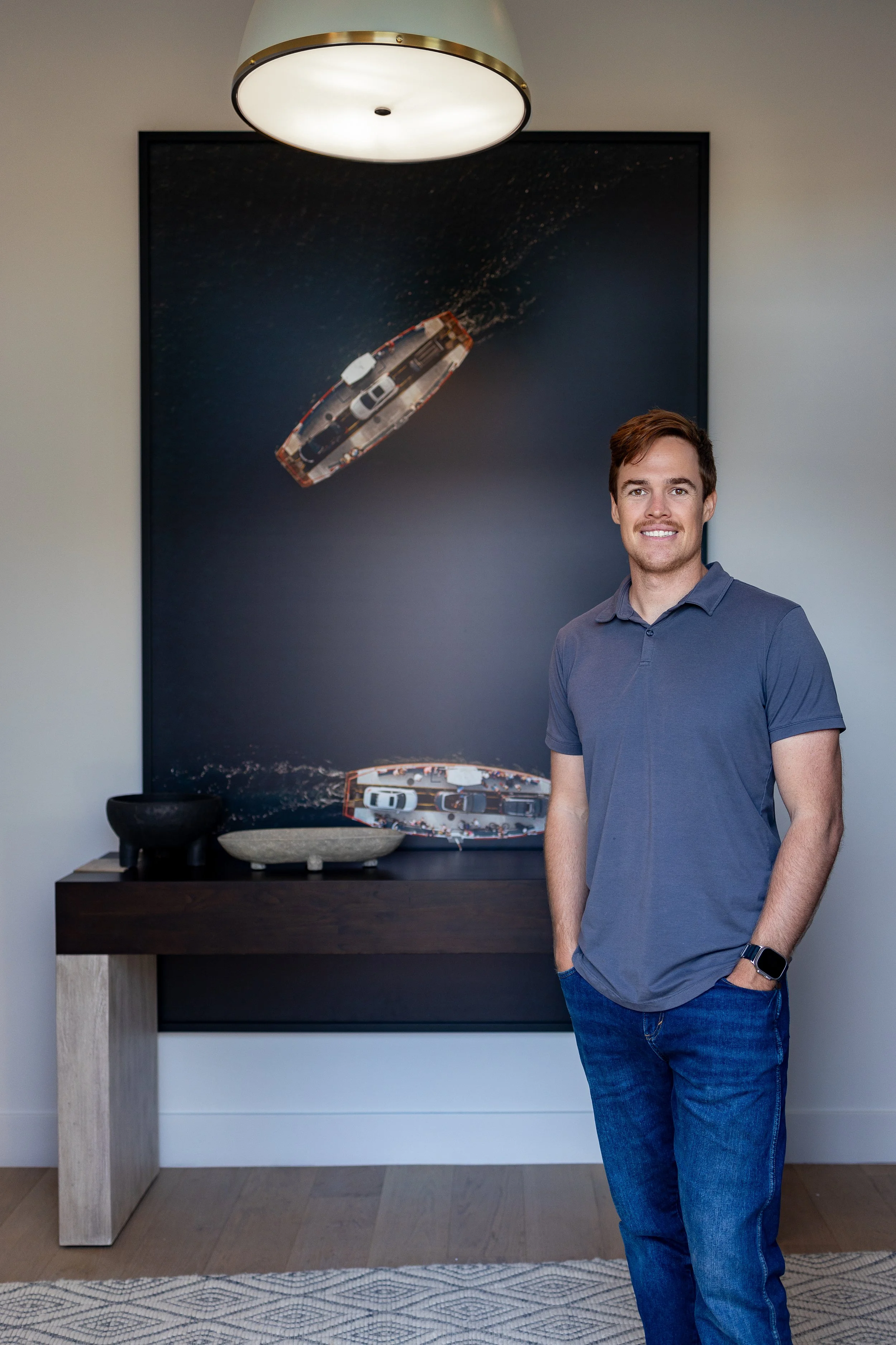 A young man with brown hair wearing a gray polo shirt and jeans, standing indoors in front of a large framed aerial photograph of boats on water, mounted on the wall.