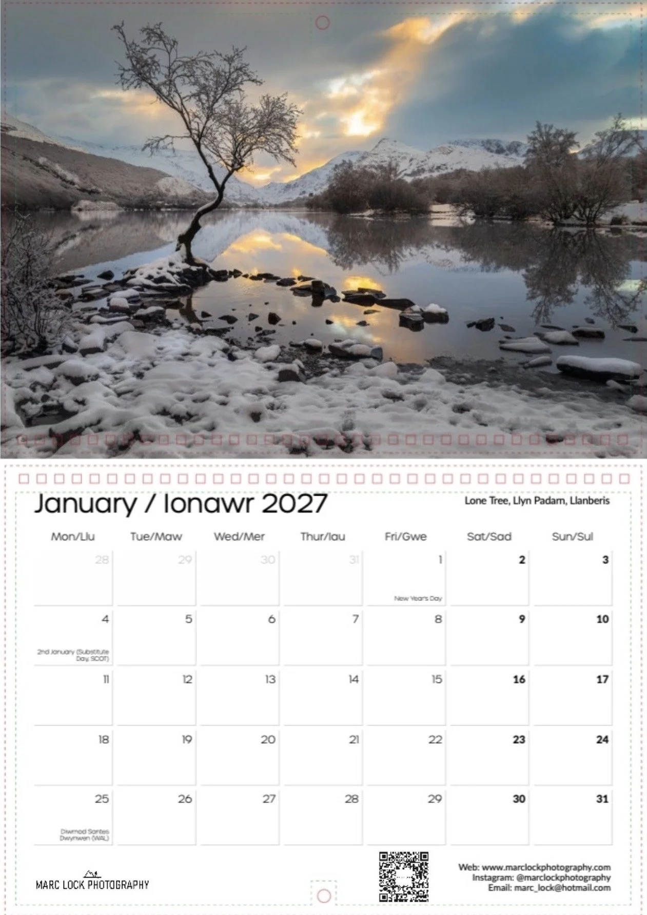 A scenic winter landscape with a lone tree by a calm lake, snow on the ground and trees, mountains in the background, and a colorful sunset sky reflected in the water.
