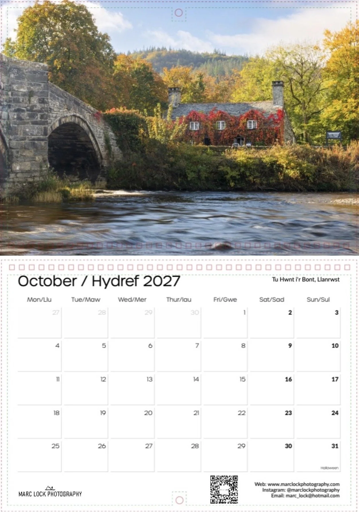A scenic photograph of a stone bridge over a river with trees and a house covered in red and green vines in the background during autumn. The sky is partly cloudy with mountains in the distance.