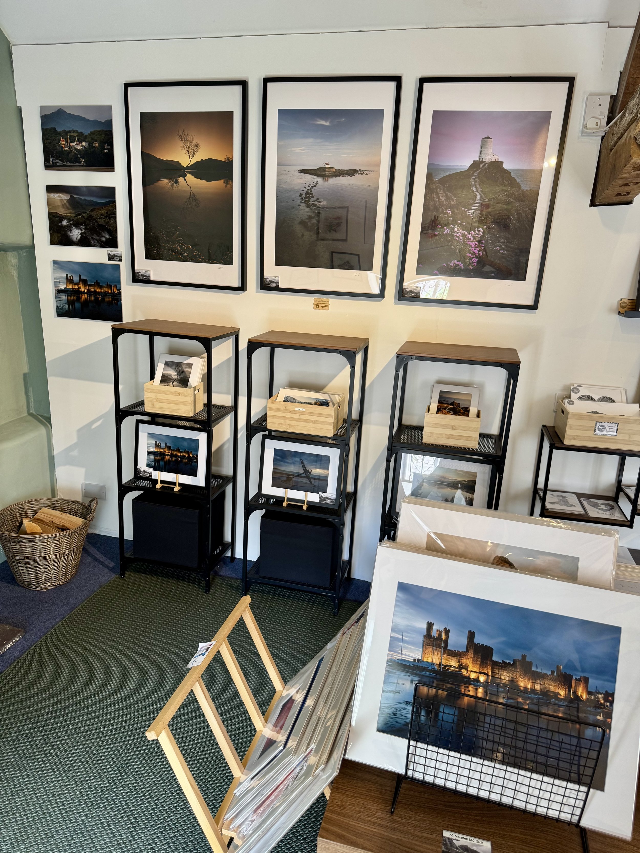 A small art gallery displays framed landscape photographs on a white wall, with additional prints in wooden and metal racks, including sunset, city, and nature scenes.