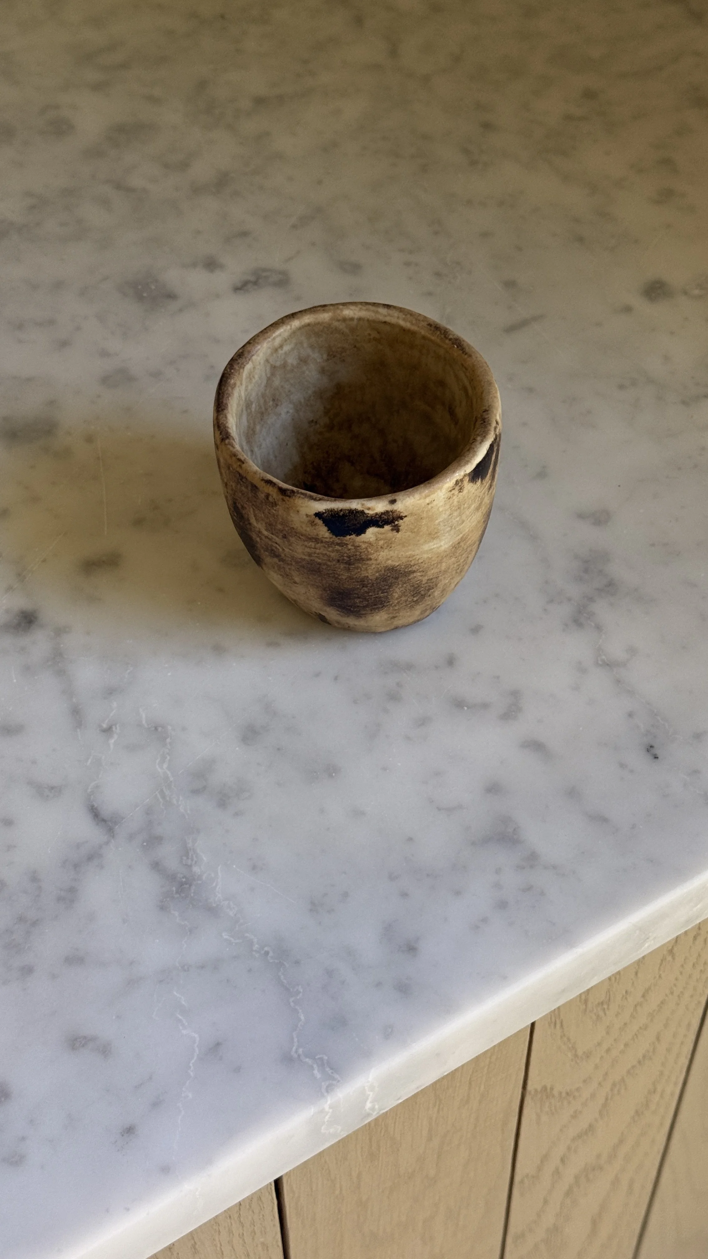 Tapered ceramic cup in warm caramel and sand tones with dark smoky patches on the surface and a pale interior