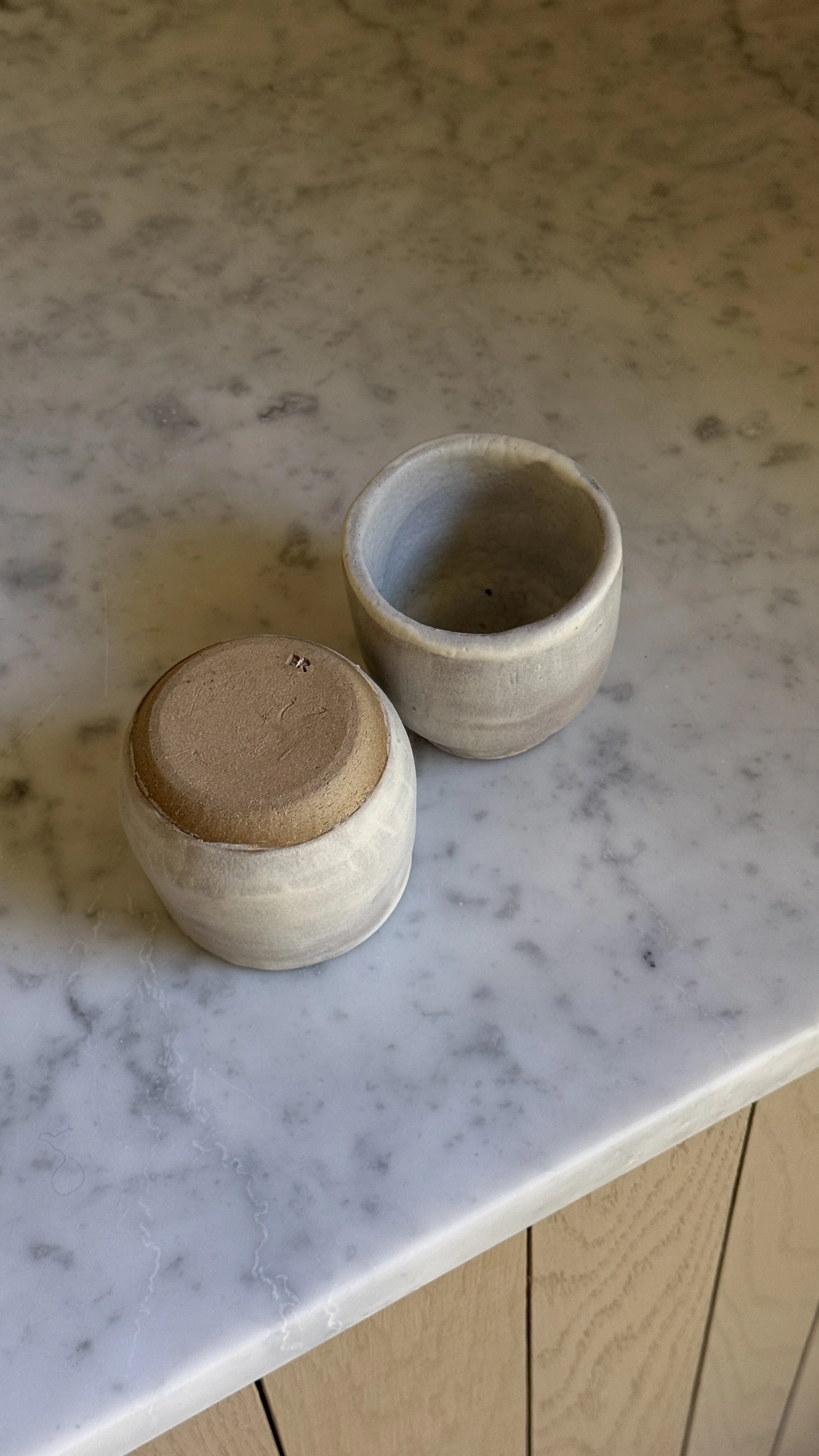 Set of two small handmade ceramic cups in a soft grey-lilac matte glaze with raw clay feet, one rounded and one straight-sided