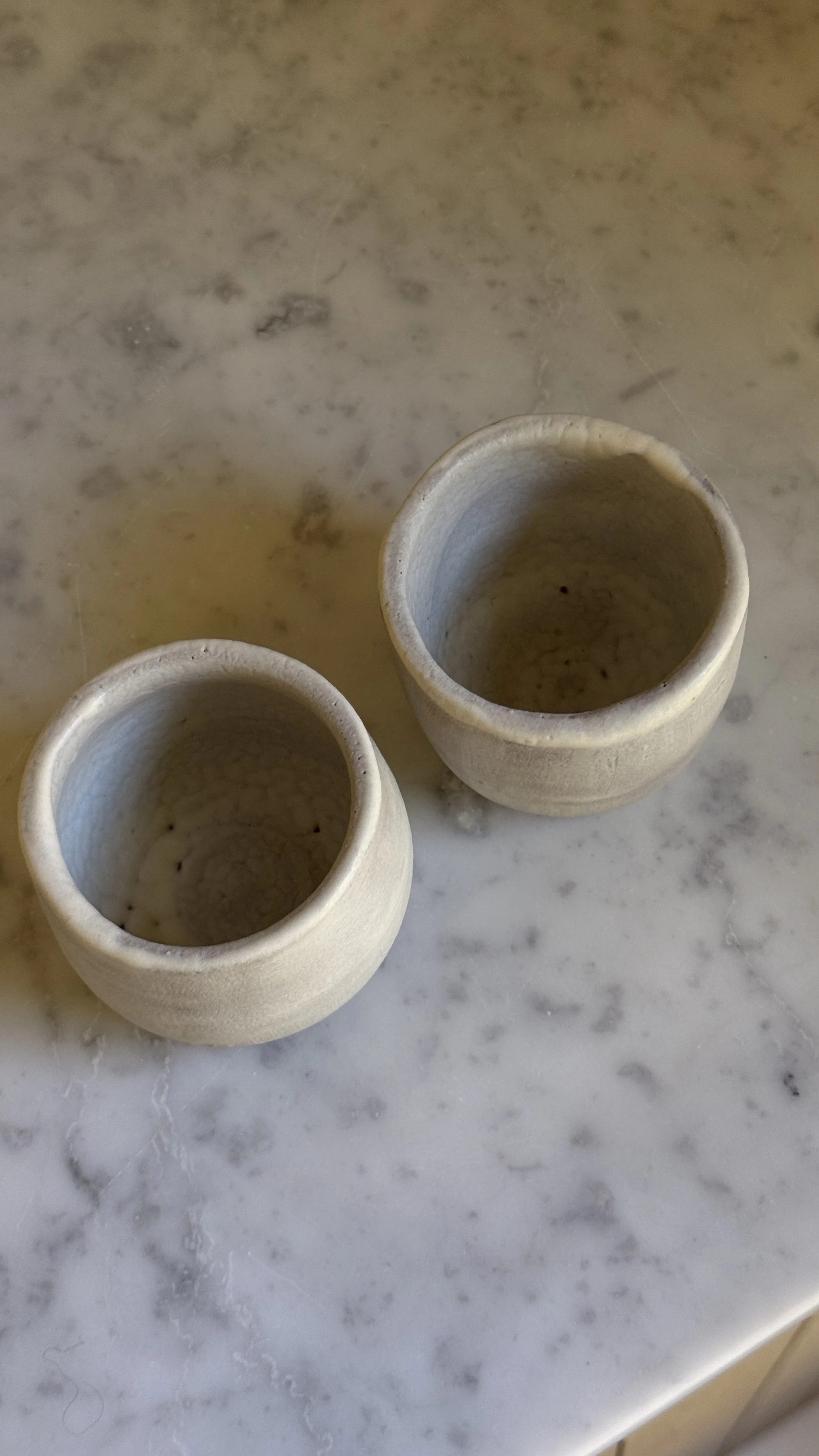 Set of two small handmade ceramic cups in a soft grey-lilac matte glaze with raw clay feet, one rounded and one straight-sided