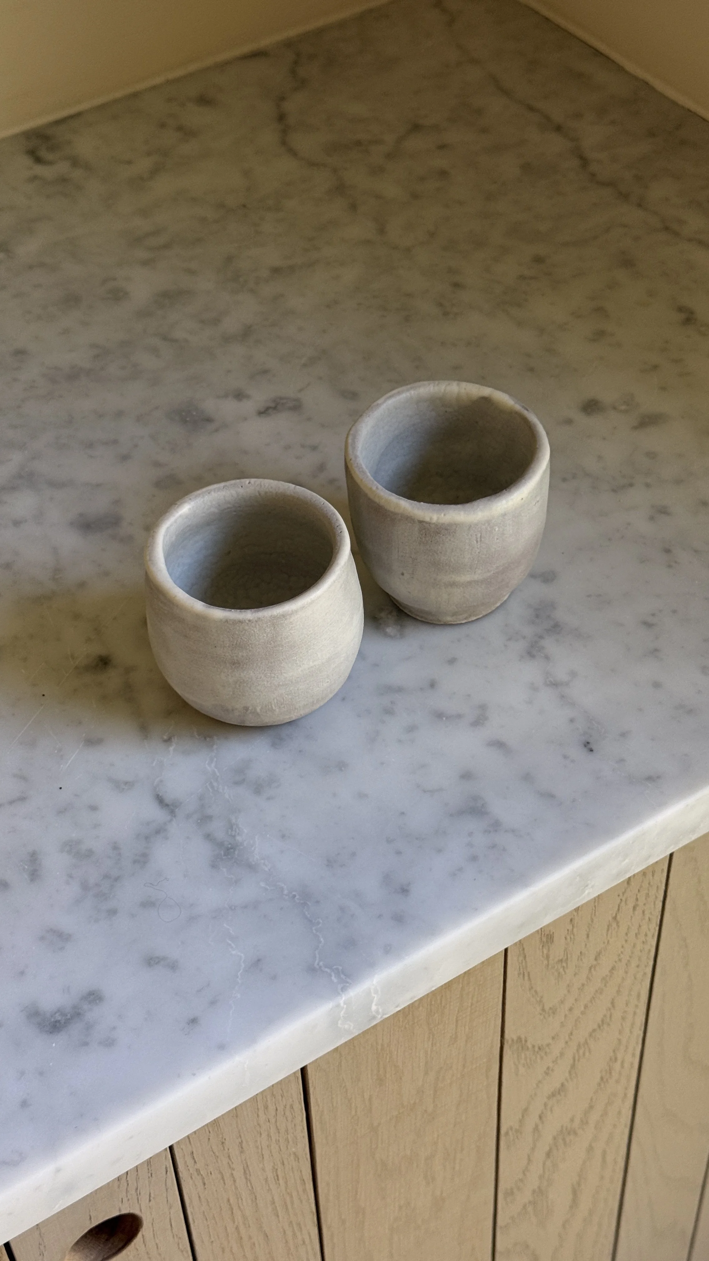 Set of two small handmade ceramic cups in a soft grey-lilac matte glaze with raw clay feet, one rounded and one straight-sided