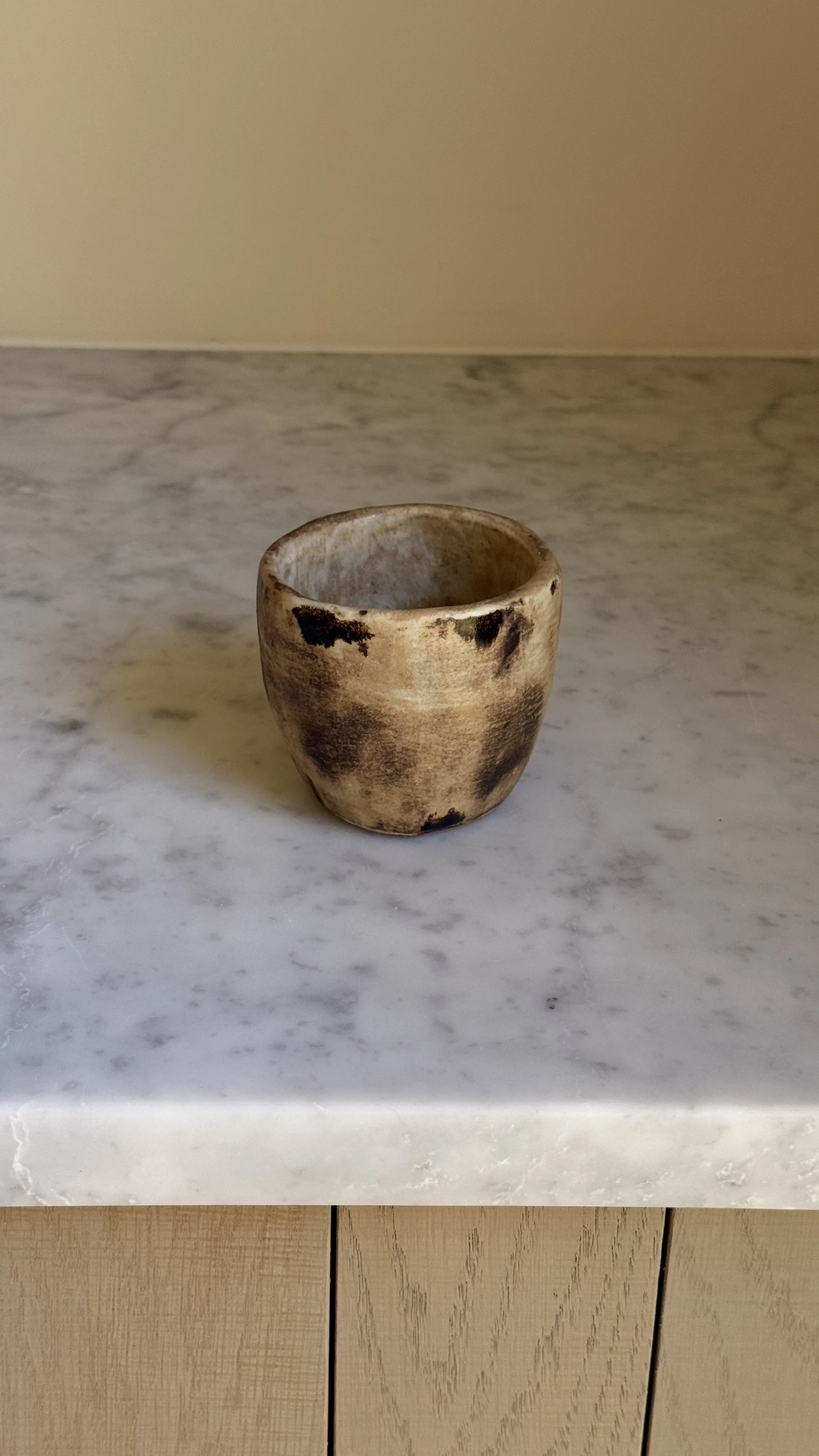 Tapered ceramic cup in warm caramel and sand tones with dark smoky patches on the surface and a pale interior