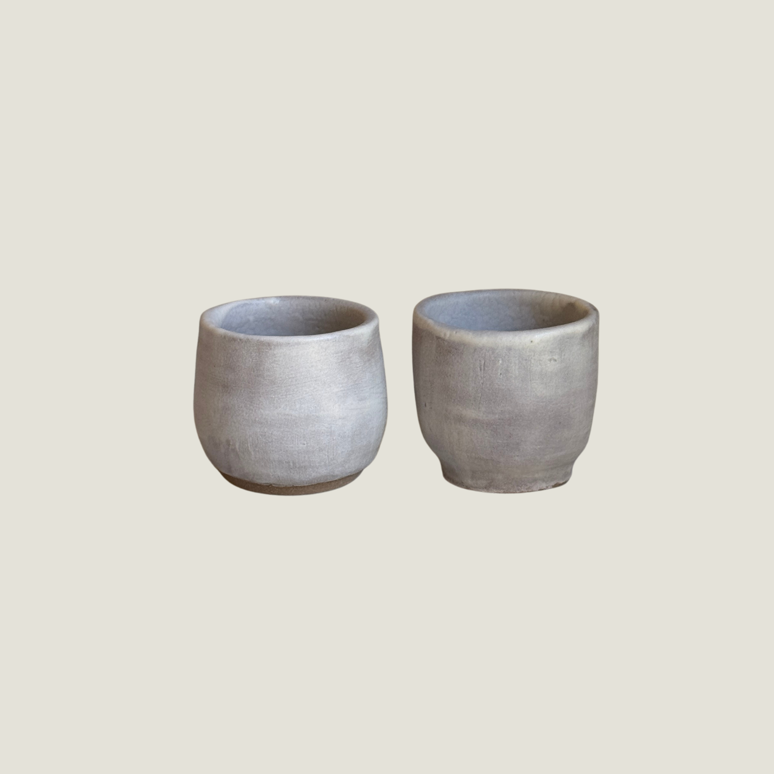 Set of two small handmade ceramic cups in a soft grey-lilac matte glaze with raw clay feet, one rounded and one straight-sided