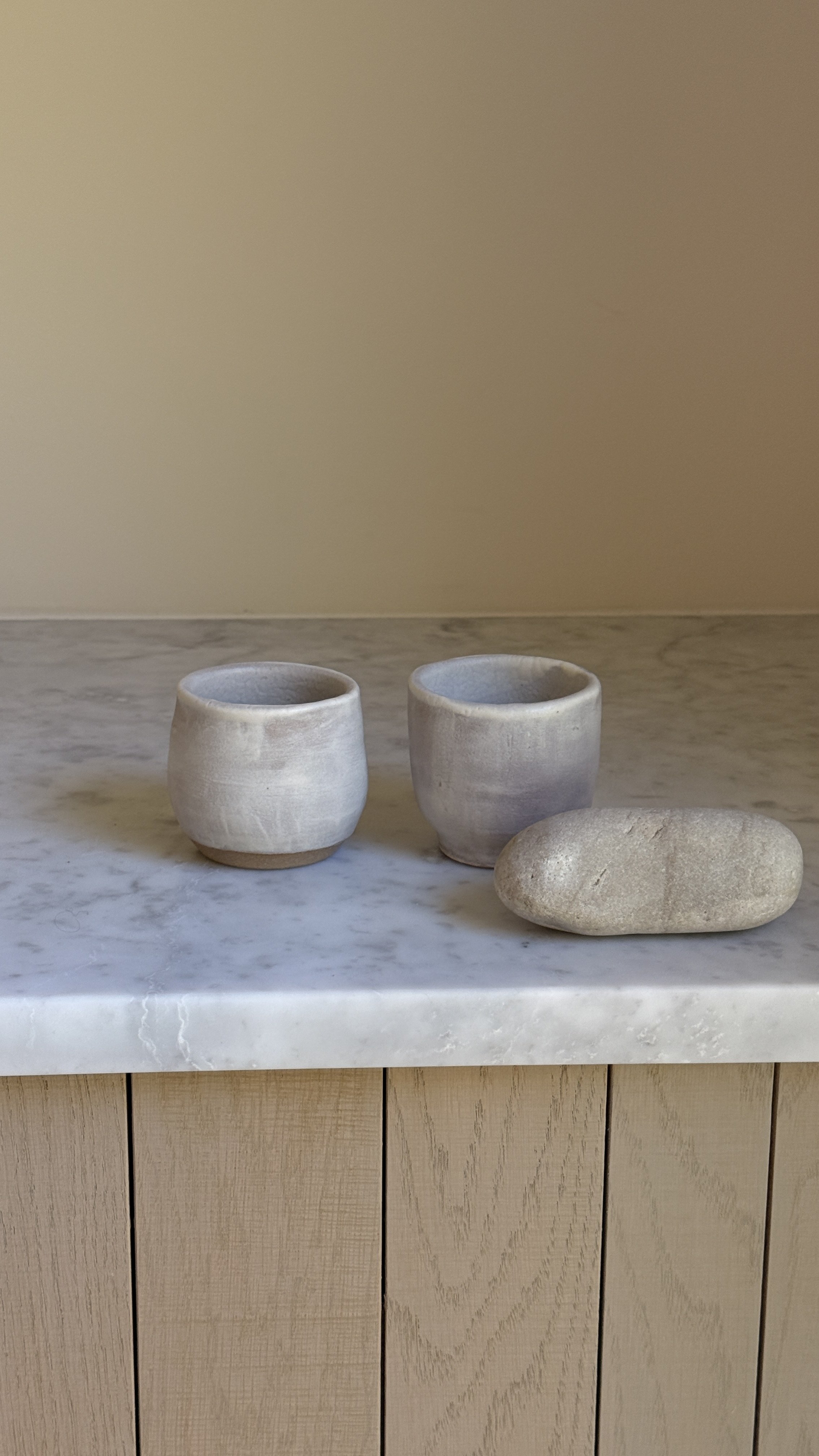 Set of two small handmade ceramic cups in a soft grey-lilac matte glaze with raw clay feet, one rounded and one straight-sided