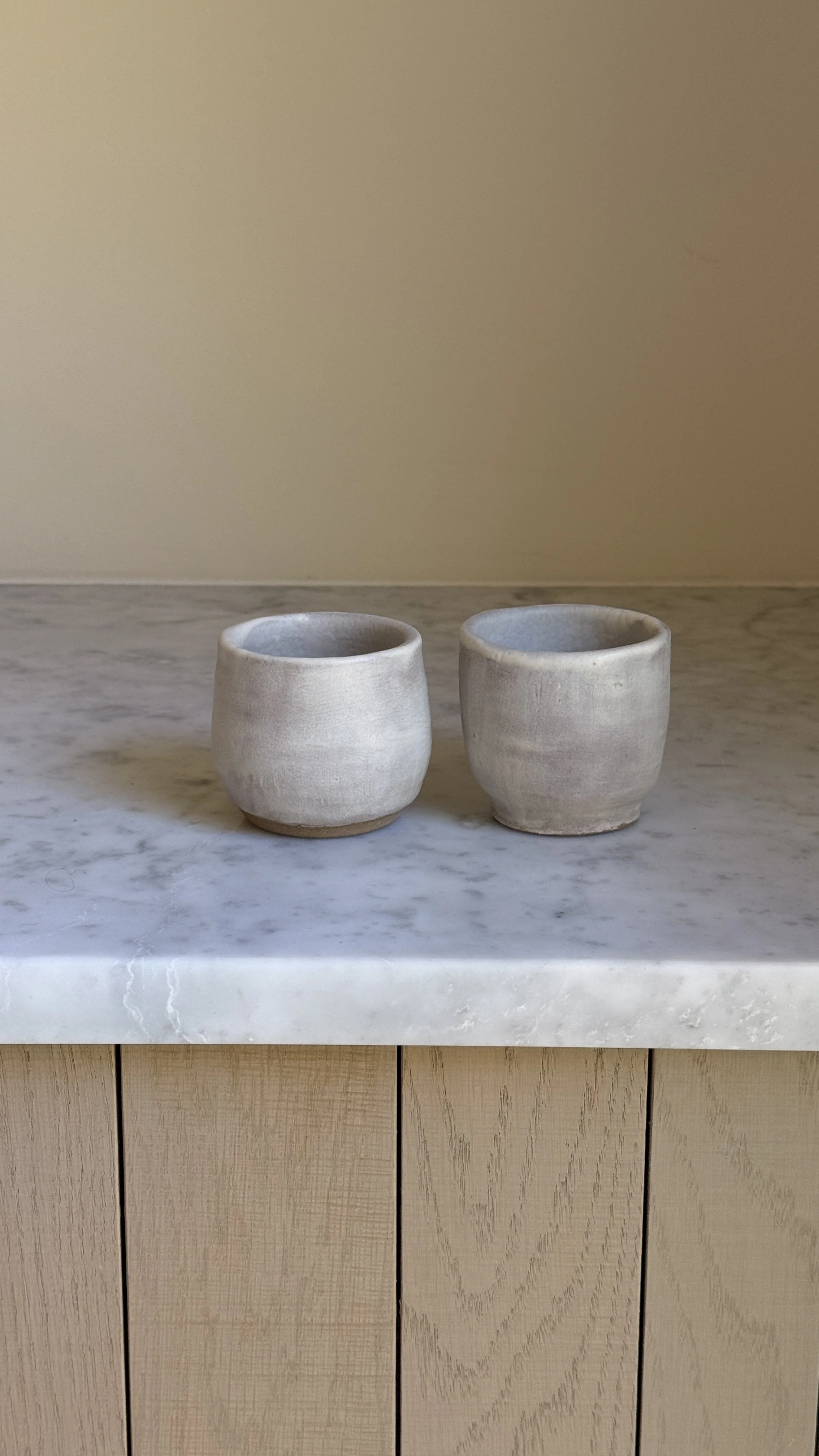 Set of two small handmade ceramic cups in a soft grey-lilac matte glaze with raw clay feet, one rounded and one straight-sided