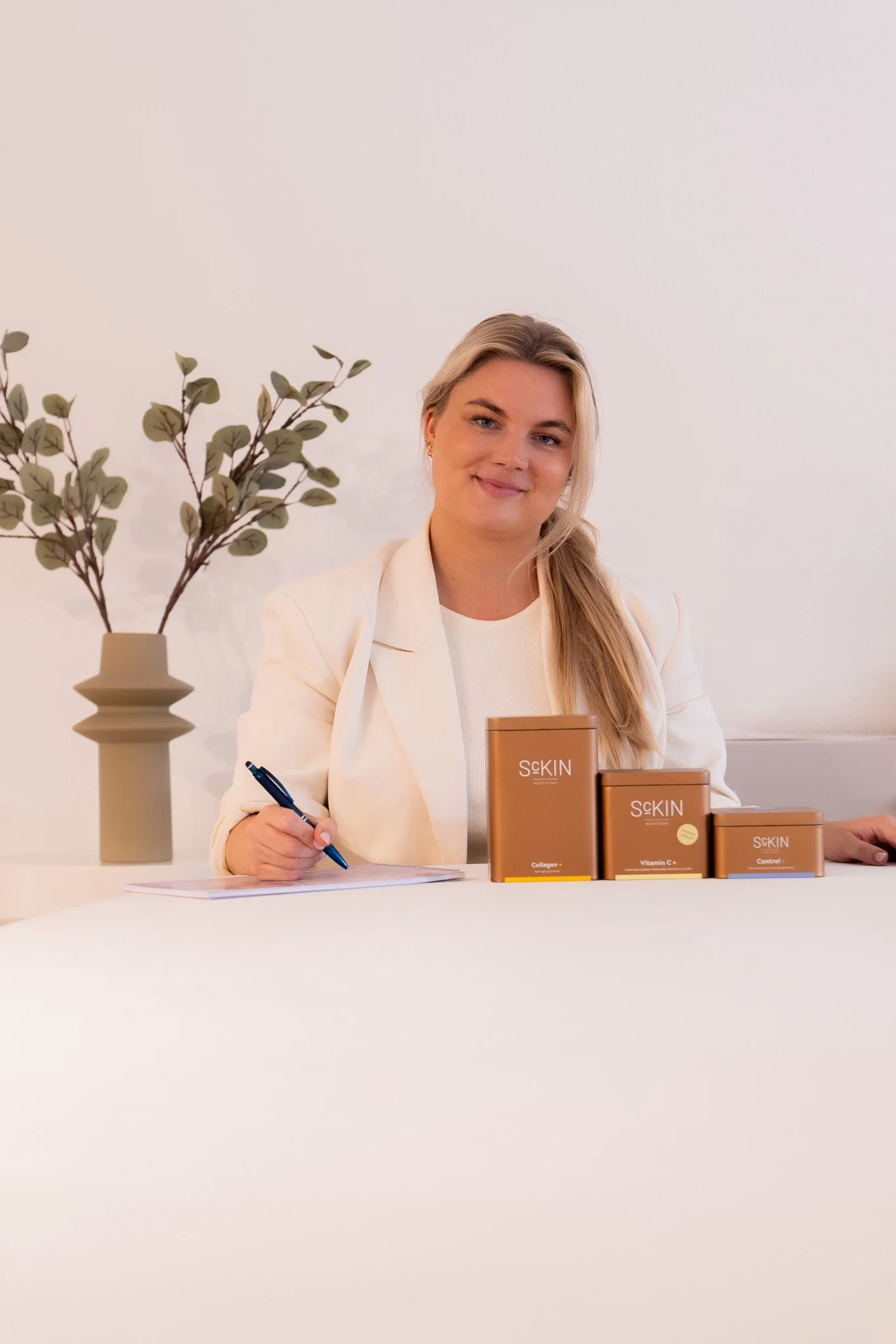 Vrouw met lange blonde haar in een witte jas en beige kleding, zittend op een lichte bank, houdt een potje met huidverzorgingsproduct, lachend naar de camera.