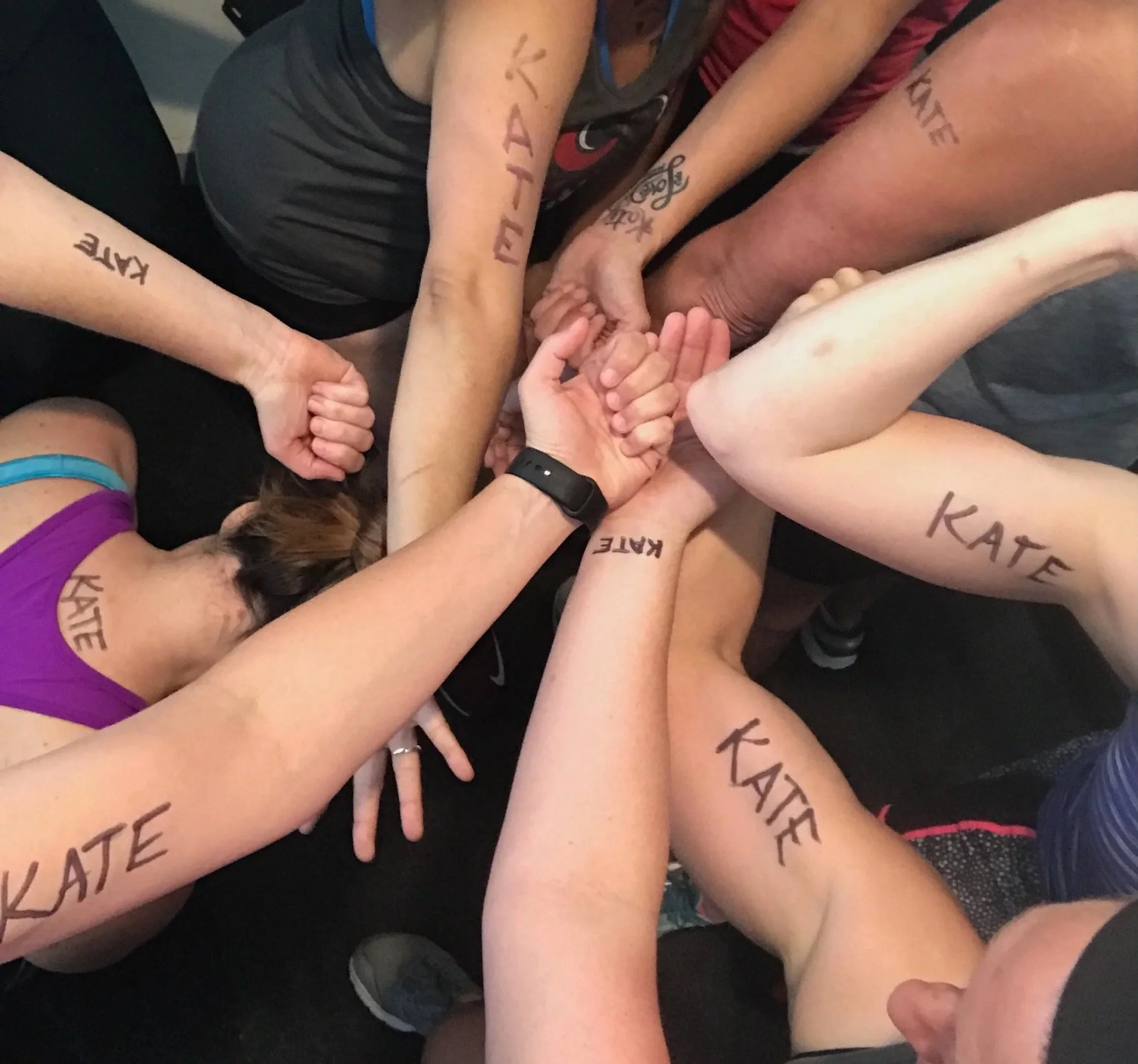 Image of a bunch of hands in a circle, many with the name Kate written on the arms
