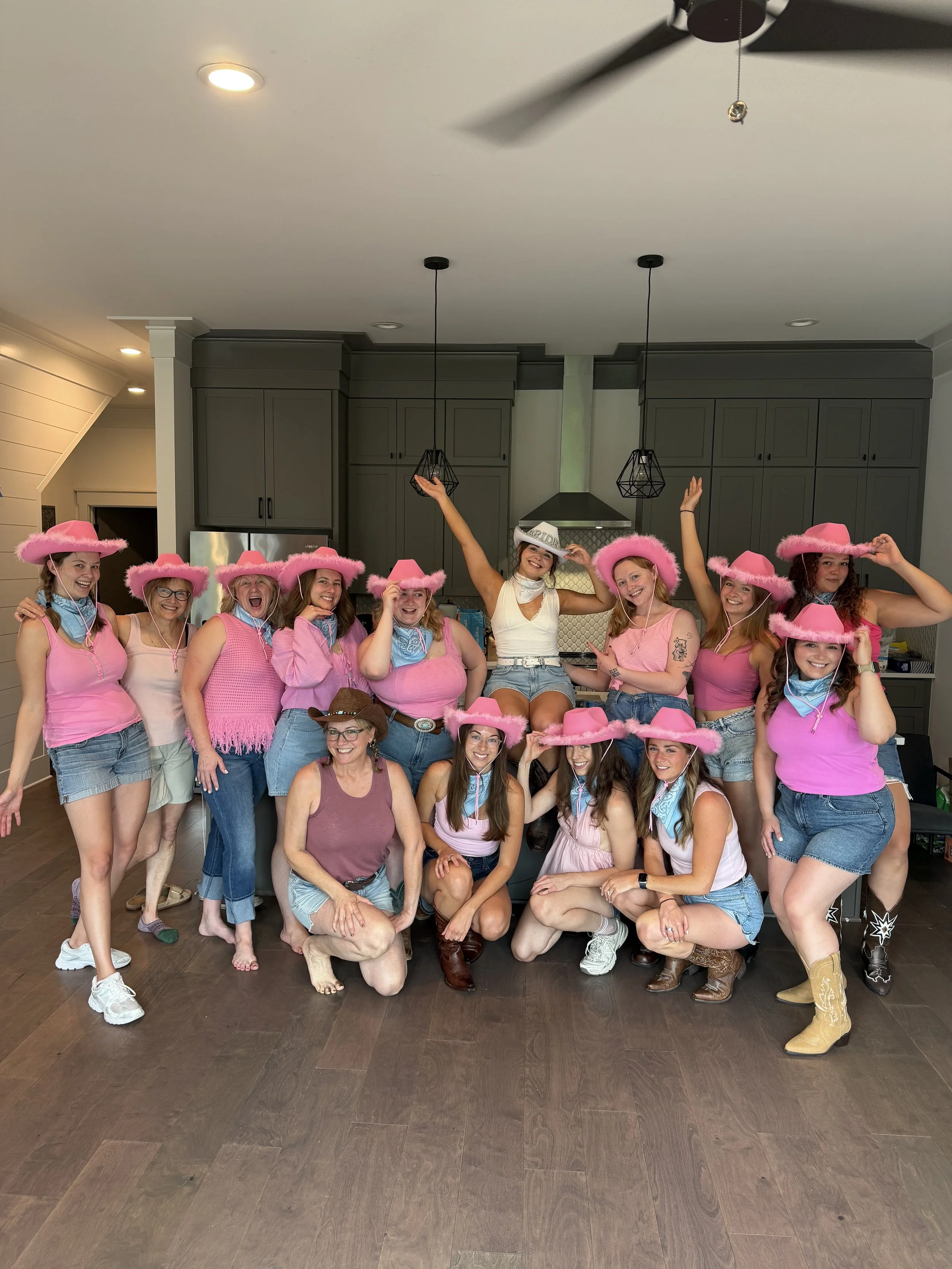 Group of women, mostly in pink cowboy hats, posing indoors in a kitchen, smiling and celebrating.