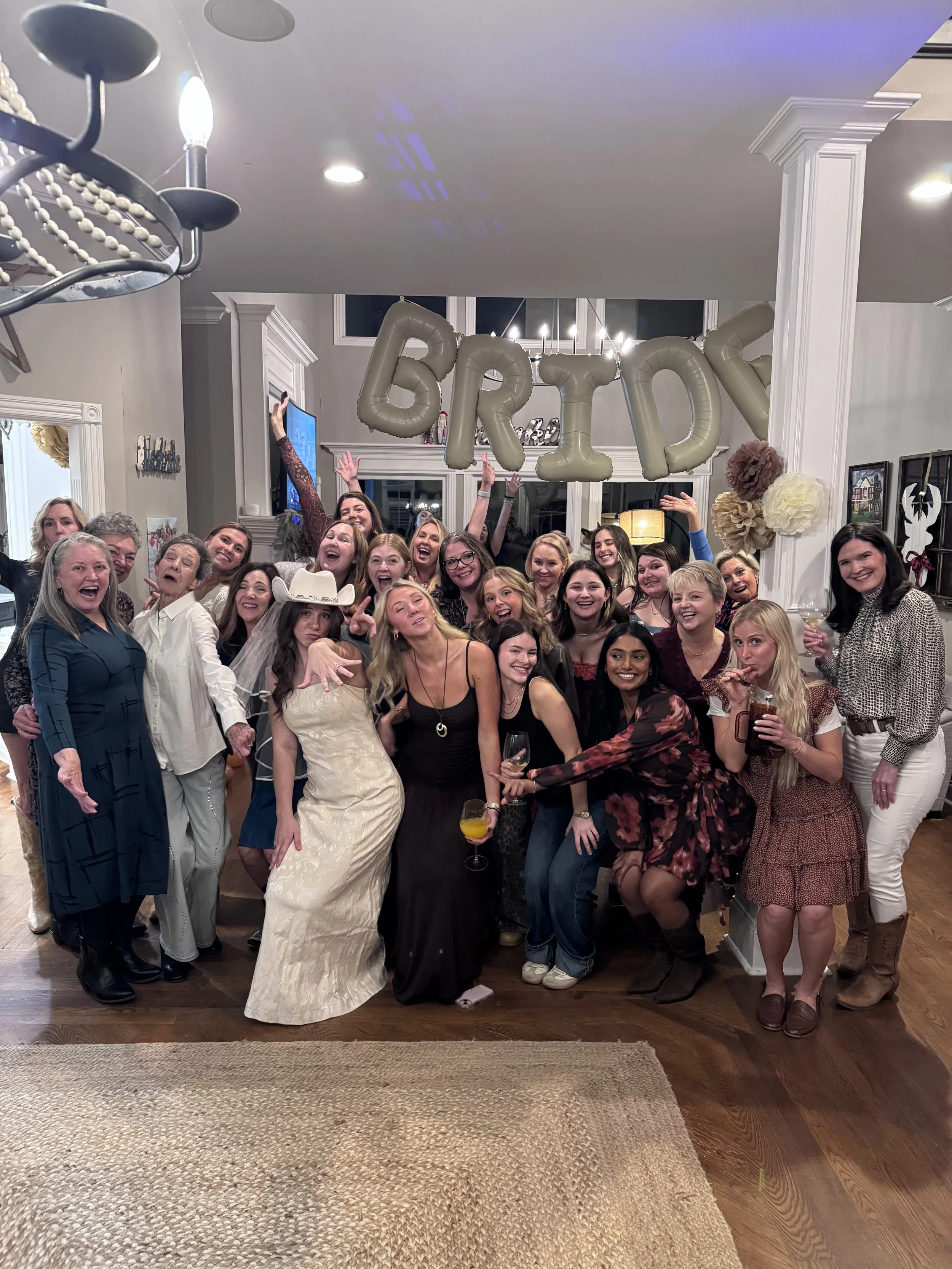 Group of women and one man celebrating at a party, with a large 'BRIDE' balloon decoration hanging in the background.