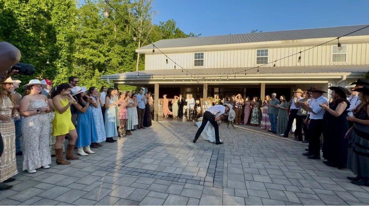 People gathered outdoors, dressed in semi-formal and cowboy attire, watching a town square dance with two men performing in the center under string lights, during daytime.