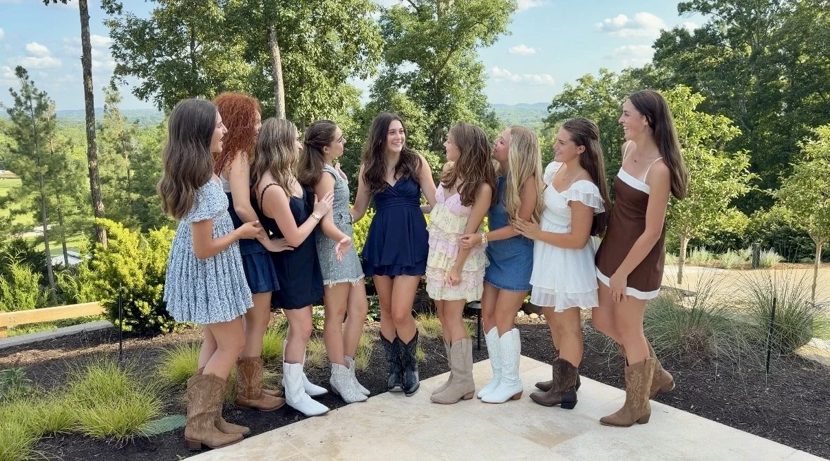 Group of nine women standing outdoors on a concrete pathway, dressed in casual summer dresses and cowboy boots, smiling and chatting in a scenic park with trees and hills in the background.