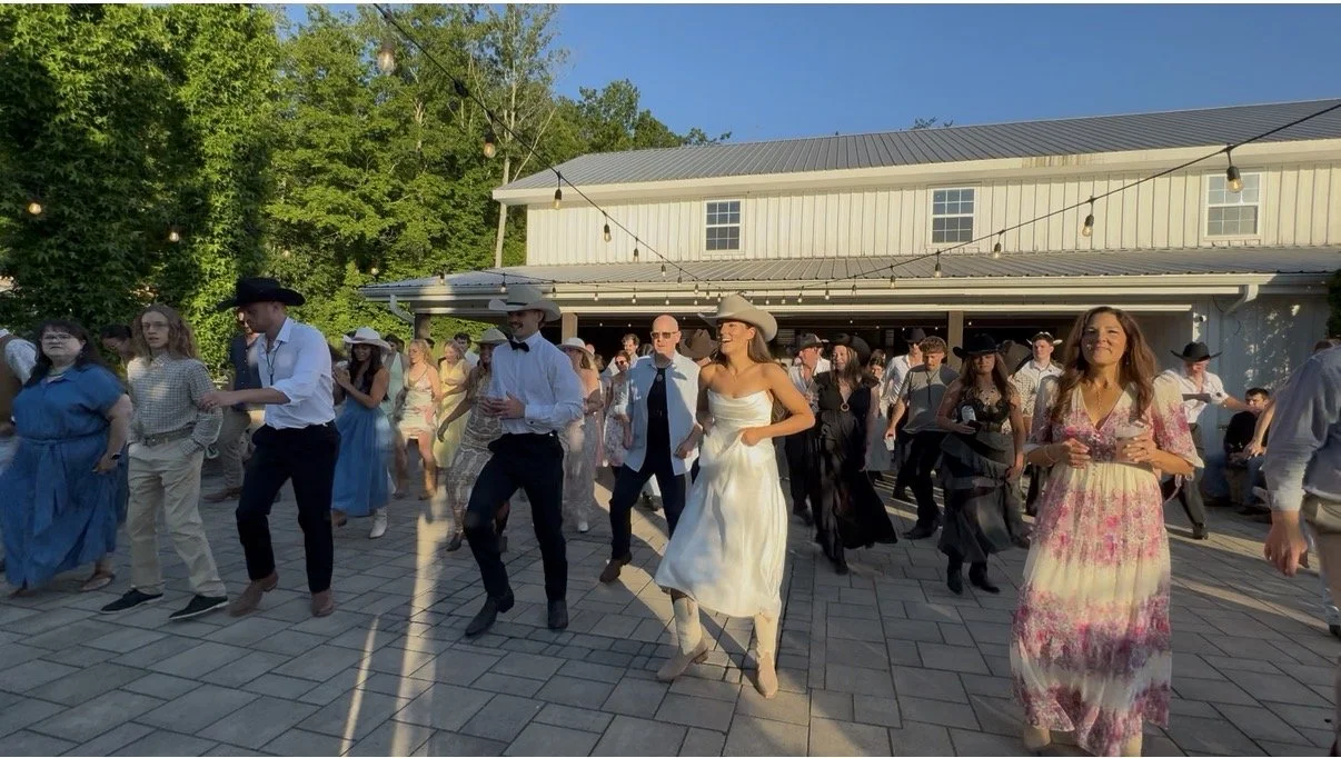People dancing outdoors during a social event at sunset, with string lights overhead and a white building in the background.