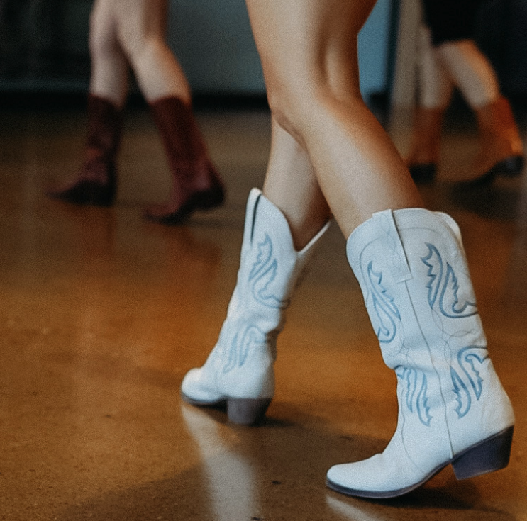 A person wearing white cowboy boots with blue embroidery, standing on a wooden floor.