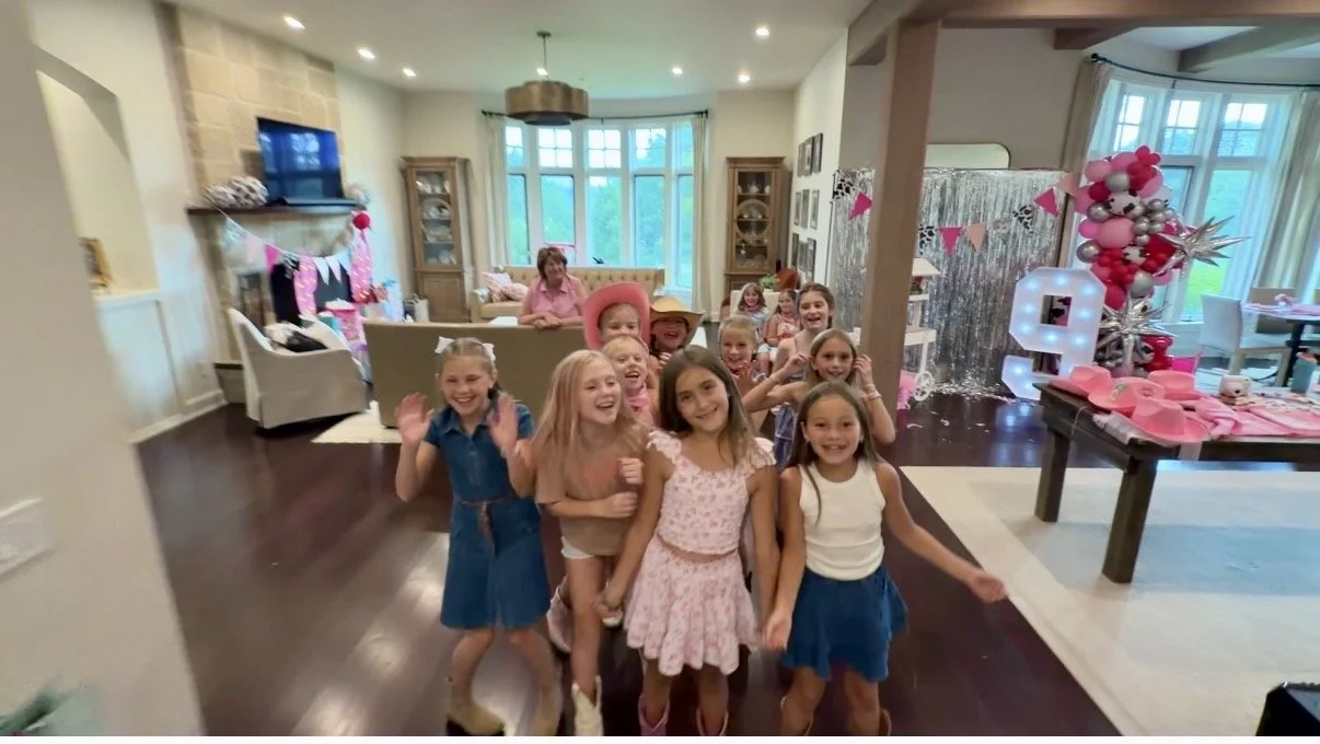 Children at a birthday party in a decorated living room with pink and silver balloons, banners, and a lit up number 6.