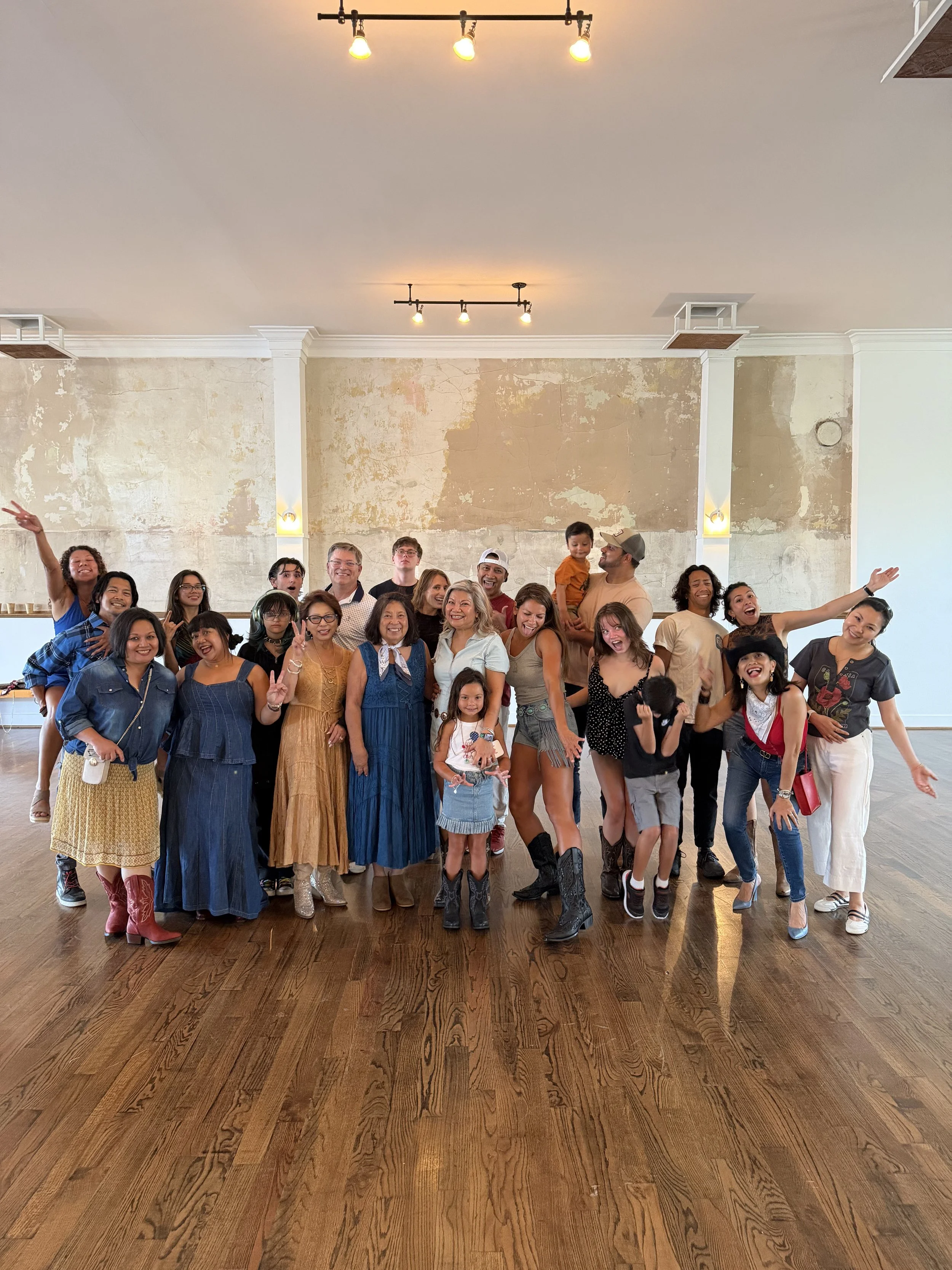 A large group of diverse people, including children and adults, is gathered together indoors, smiling and posing for a photo on a wooden floor with a textured wall and ceiling lights in the background.