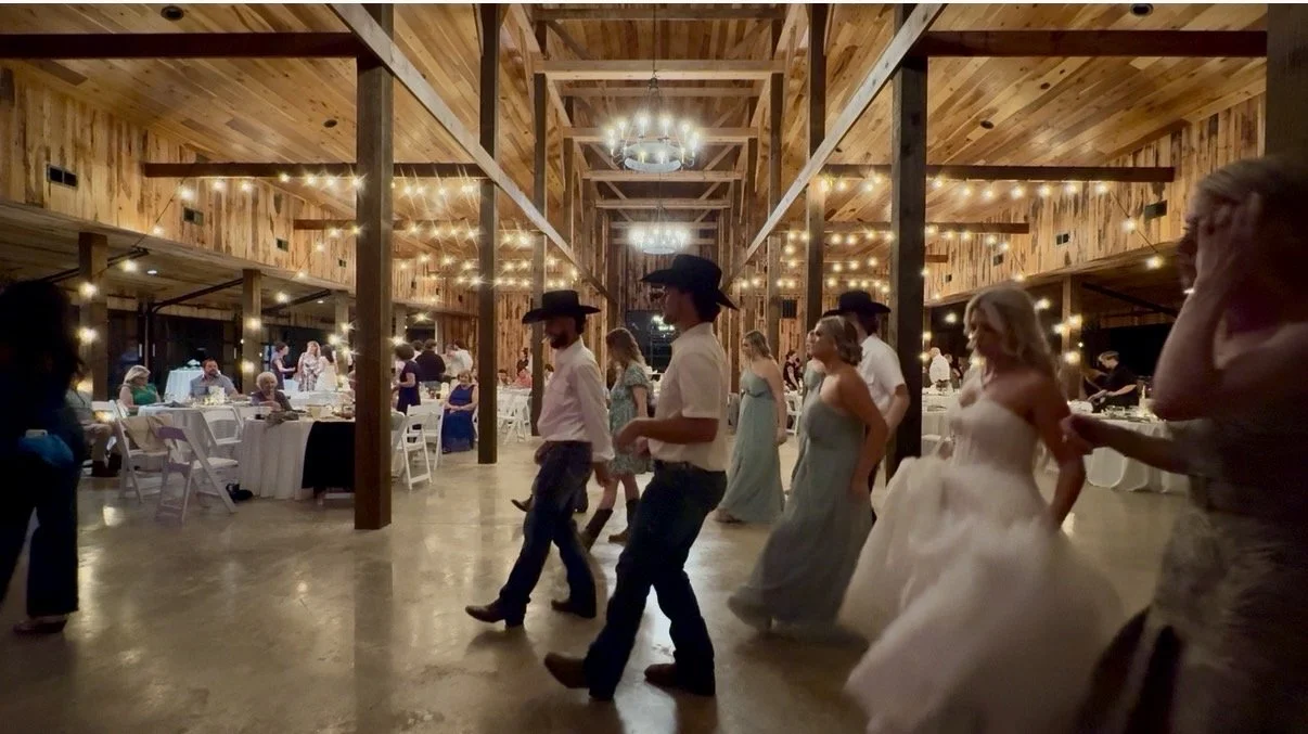 Guests dancing at a wedding reception inside a rustic wooden hall decorated with string lights.