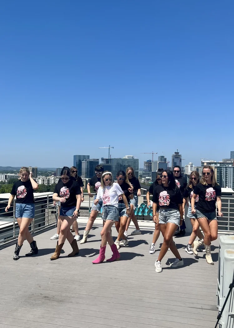 A group of young women dancing on a rooftop with a city skyline in the background under a clear blue sky.