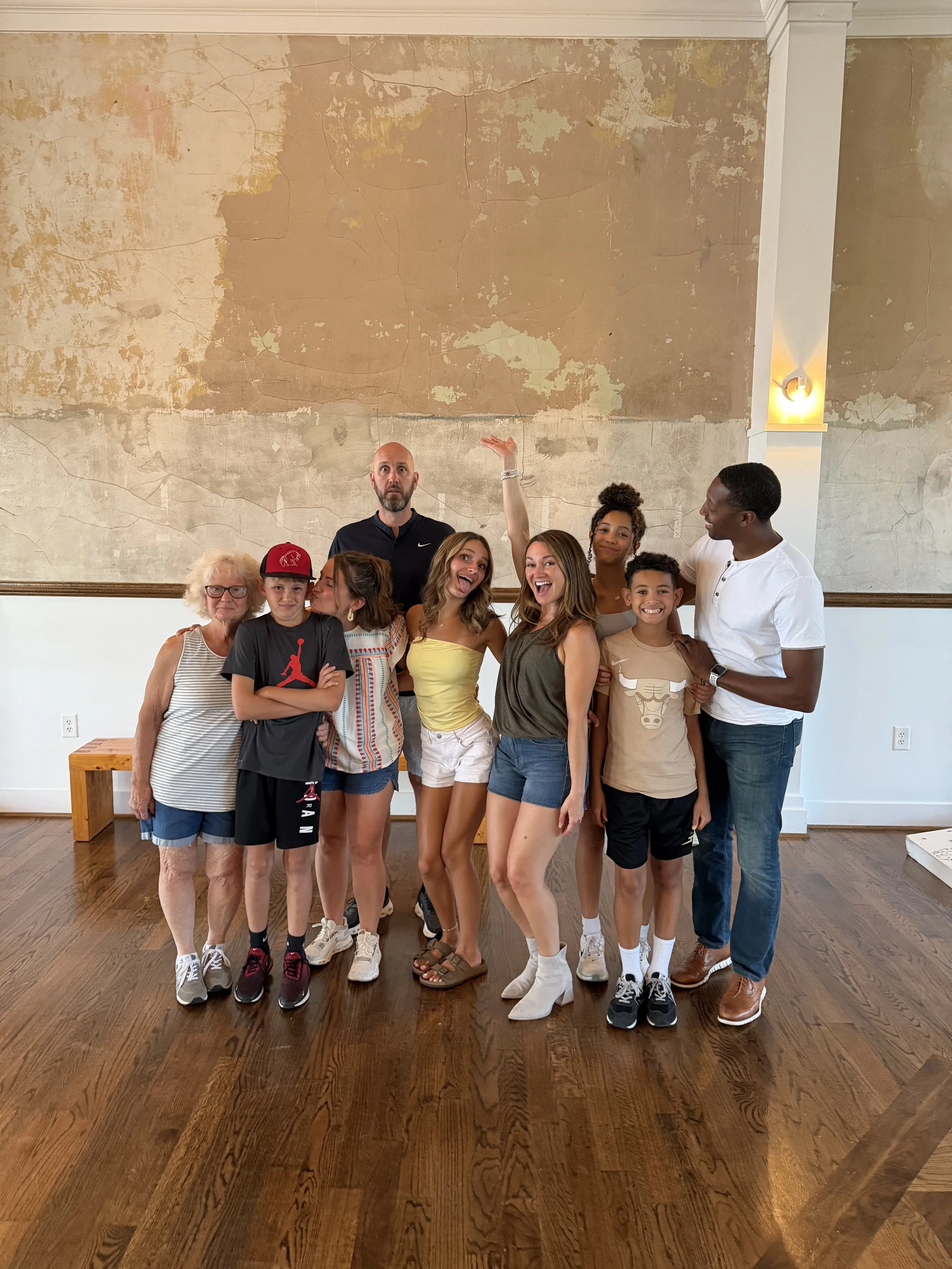 A group of ten people, including children and adults, posing together indoors in front of a textured, cracked wall with a white column and a wall light fixture. They are smiling, with one woman raising her arm and a man looking at the camera.