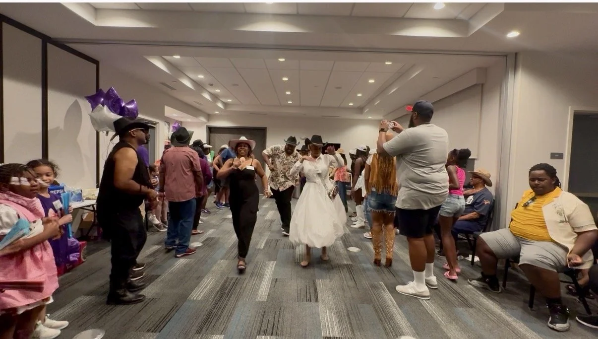 People dancing in a decorated indoor party room with several children and adults, some wearing cowboy hats, and balloons on the side.