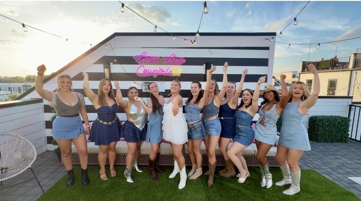 Group of women celebrating on a rooftop patio at sunset, some with their arms raised, with a neon sign that says "Lonny-Luxe Cougars" in the background.