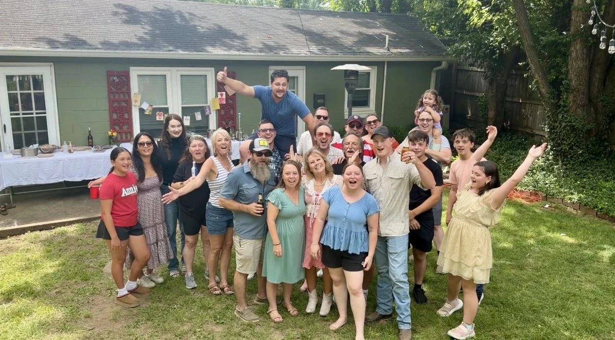 Group of people celebrating in a backyard, some with drinks, with one person sitting on others' shoulders, and a table with food and drinks in the background.