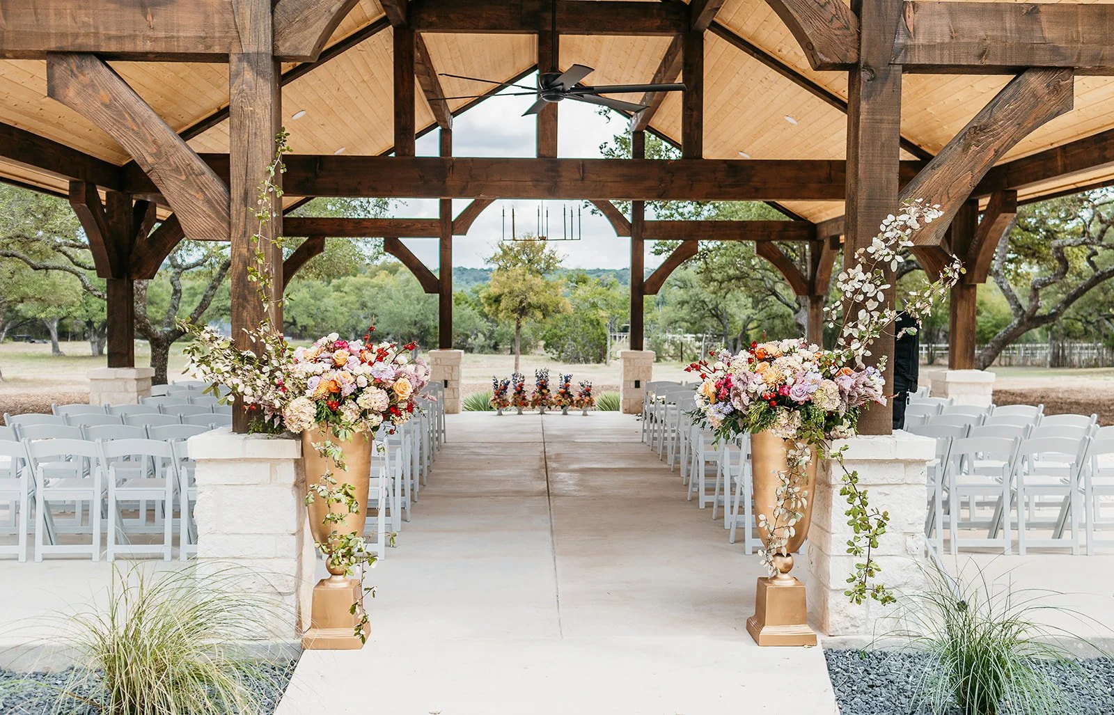 Outdoor wedding ceremony setup under a wooden pavilion with floral arrangements on pedestals at the entrance, rows of white chairs on each side, and a view of trees and hills in the background.