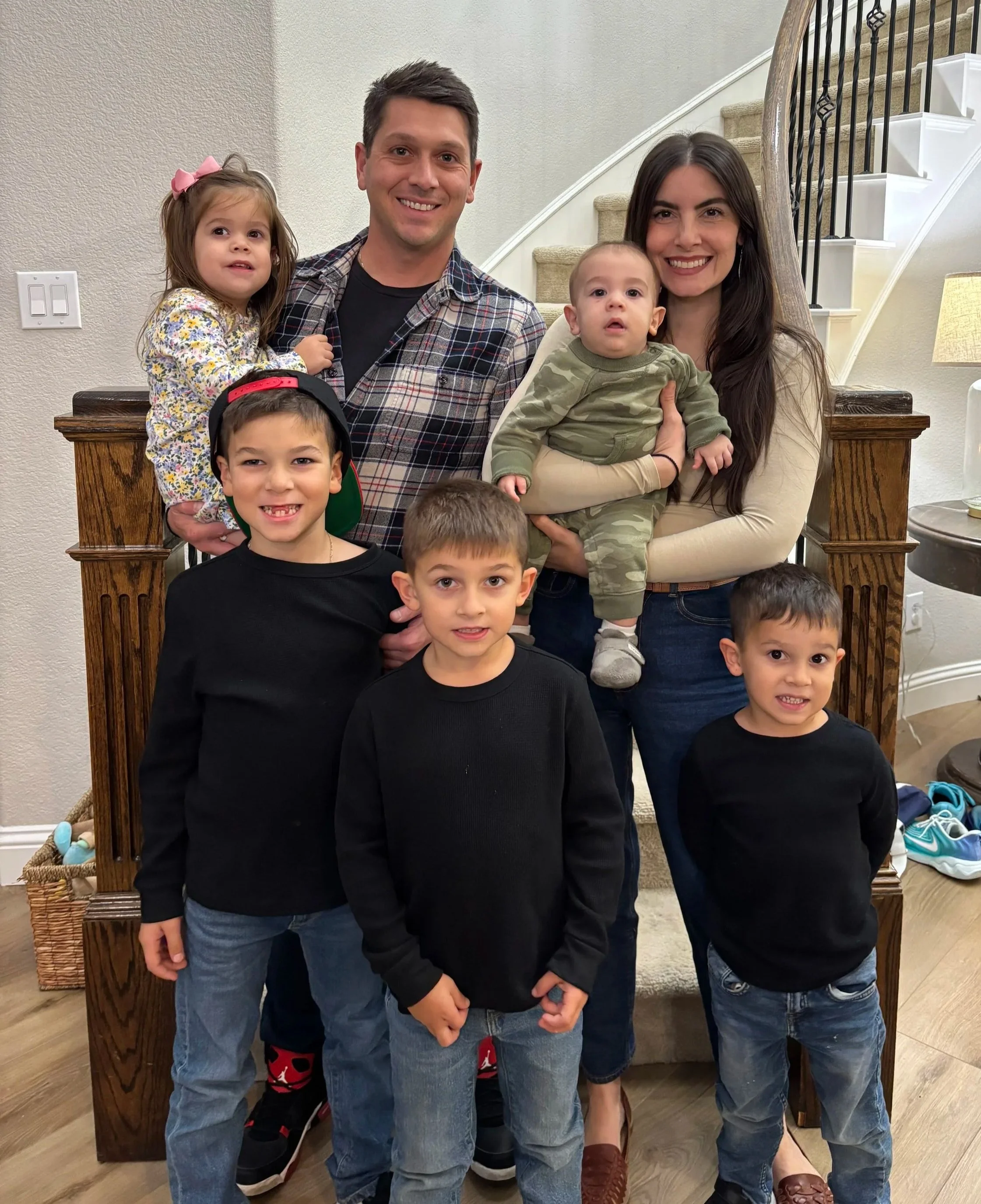 A family portrait featuring two adults and five children posing together indoors in front of a staircase.