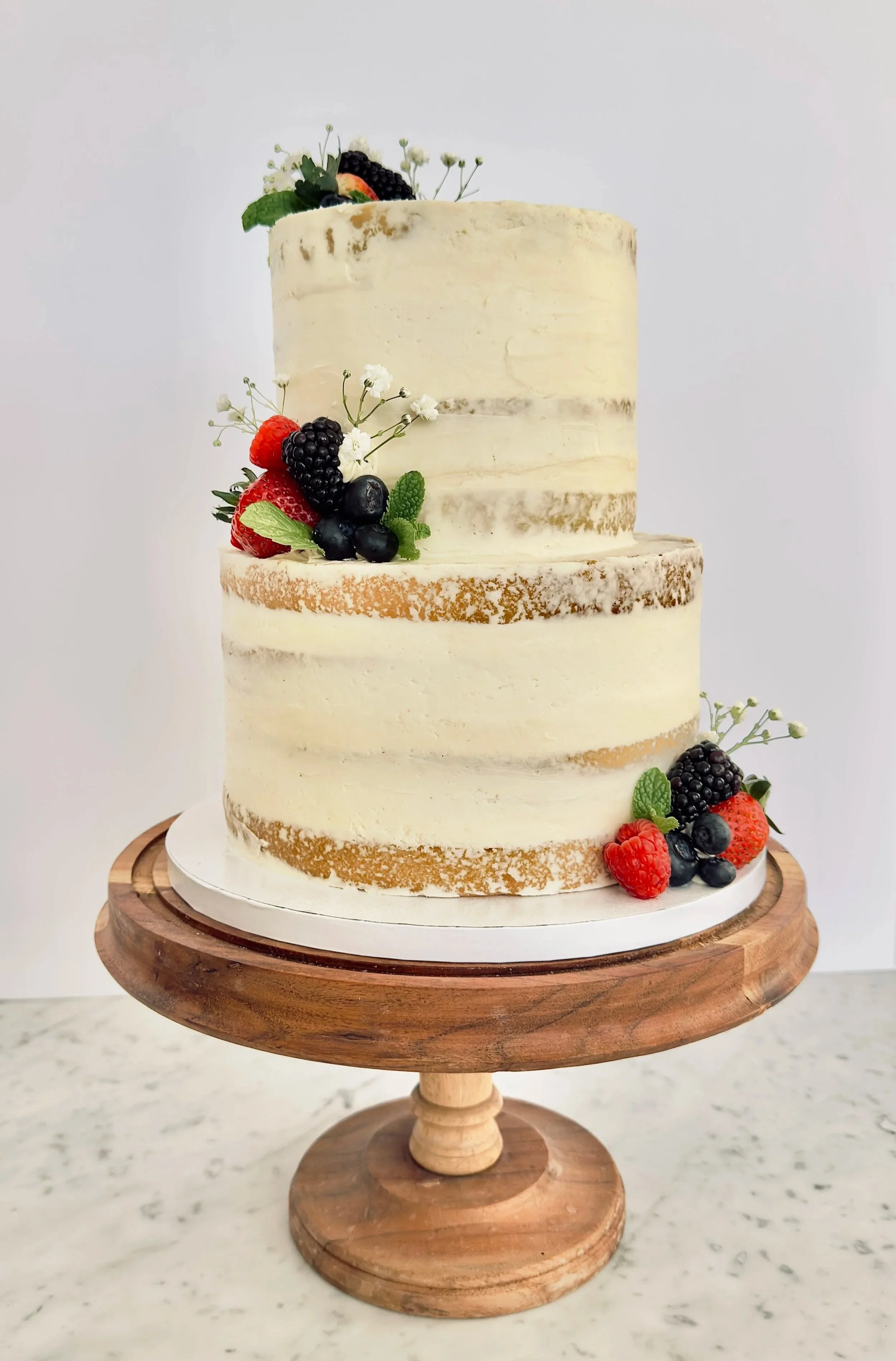 Two-tiered naked cake with white frosting, decorated with fresh berries and small white flowers, placed on a wooden cake stand.
