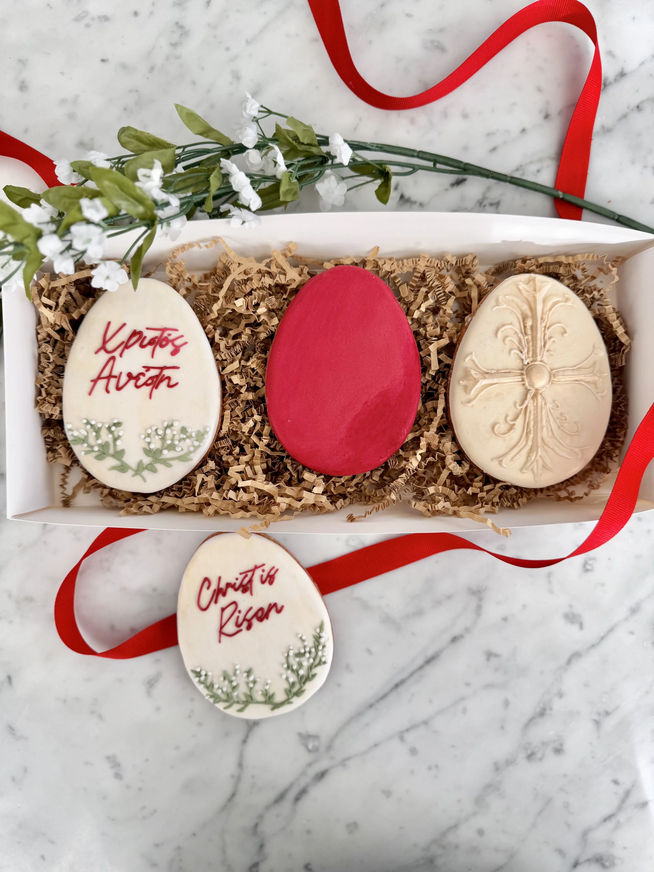 Decorated Easter eggs in a white box with crinkled paper, one with red writing and a green leaf design, another solid red, and another with ornate white patterns, on a marble surface with a red ribbon and a small sprig of white flowers.
