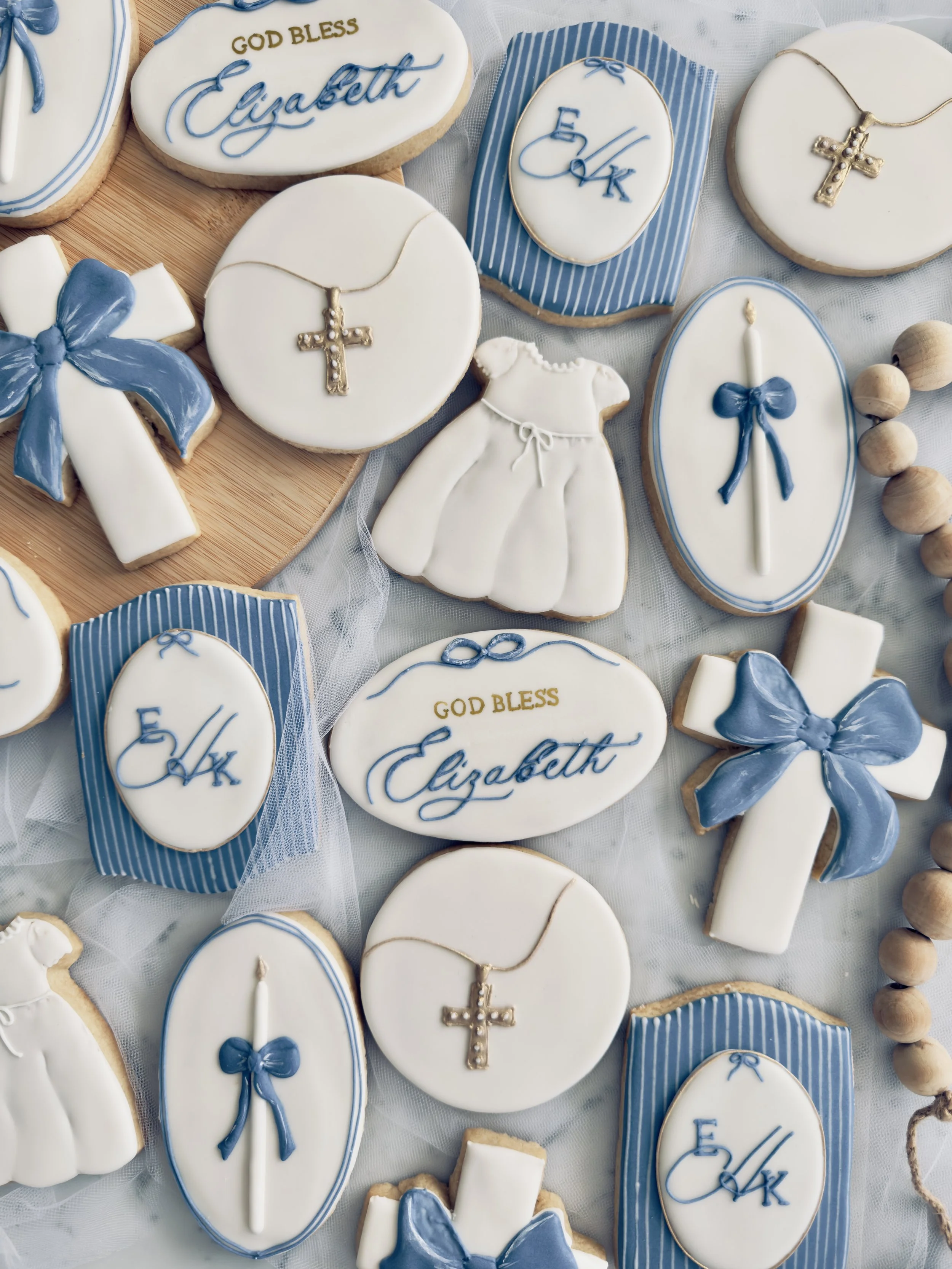 Decorated cookies with religious and baptism themes, including crosses, baptism gowns, 'God Bless Elizabeth' messages, and blue bows, arranged on a table.