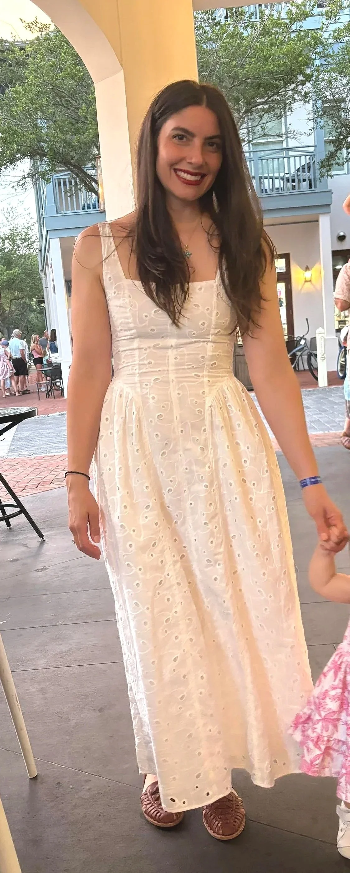 A woman with long dark hair, wearing a white sleeveless dress with eyelet embroidery, standing outdoors on a brick patio. She is smiling, holding hands with a child, and has a building with balcony and trees in the background.