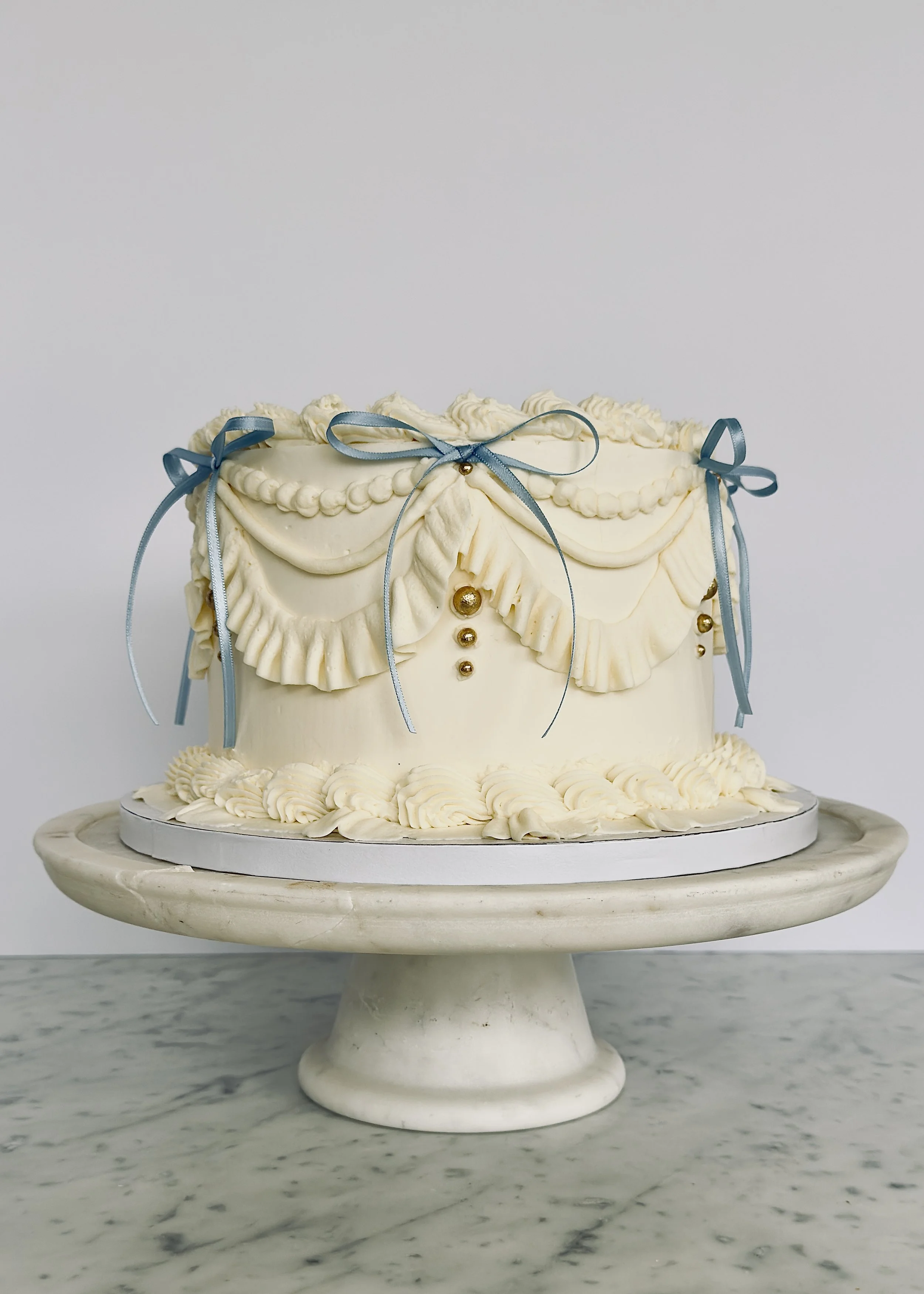 Elegant white cake with piped frosting, blue ribbons, and gold accents on a white cake stand.