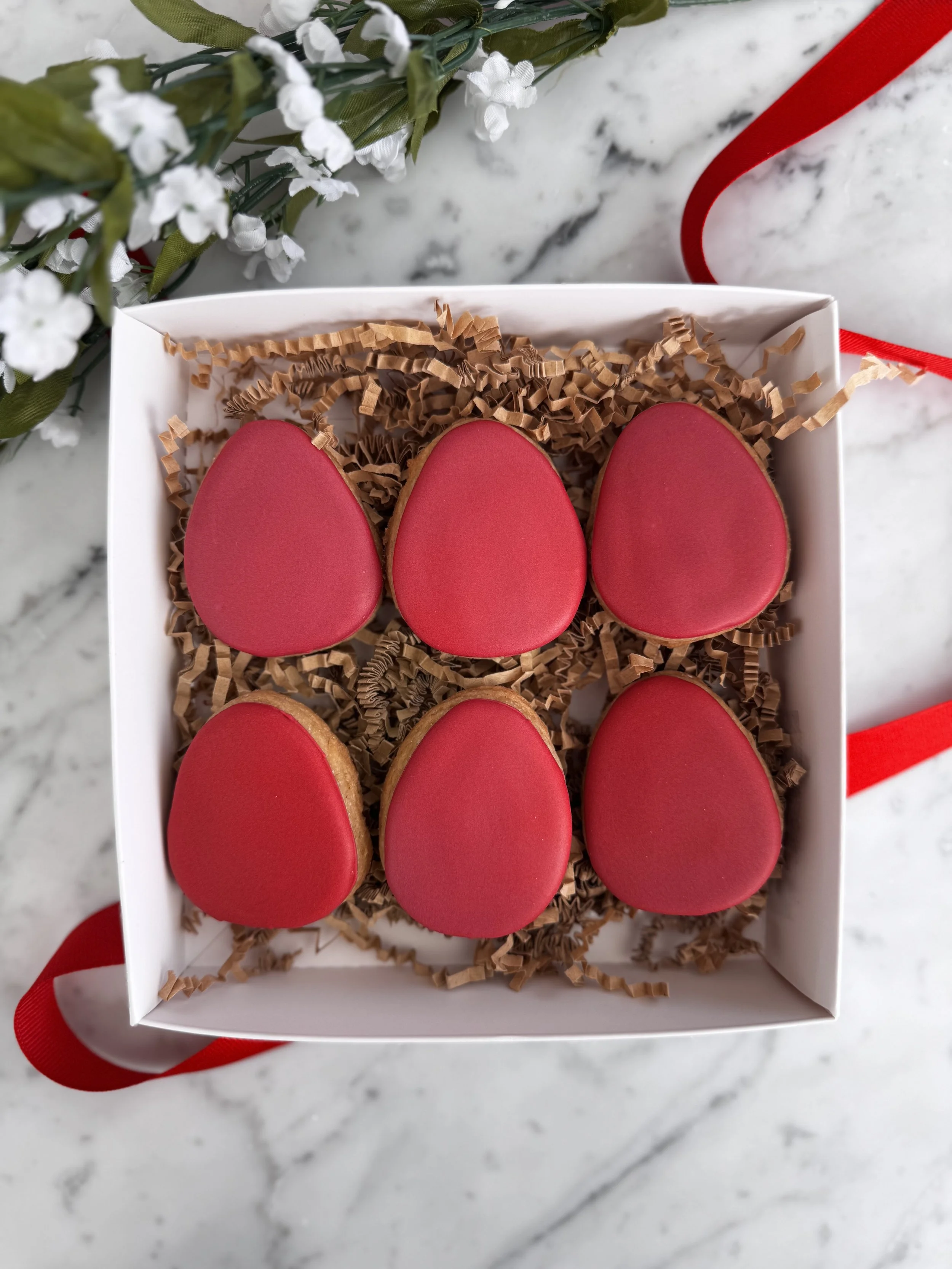 Box of six red heart-shaped cookies decorated with red icing, arranged on brown shredded paper, with a white flower and green leaves nearby on a marble surface.