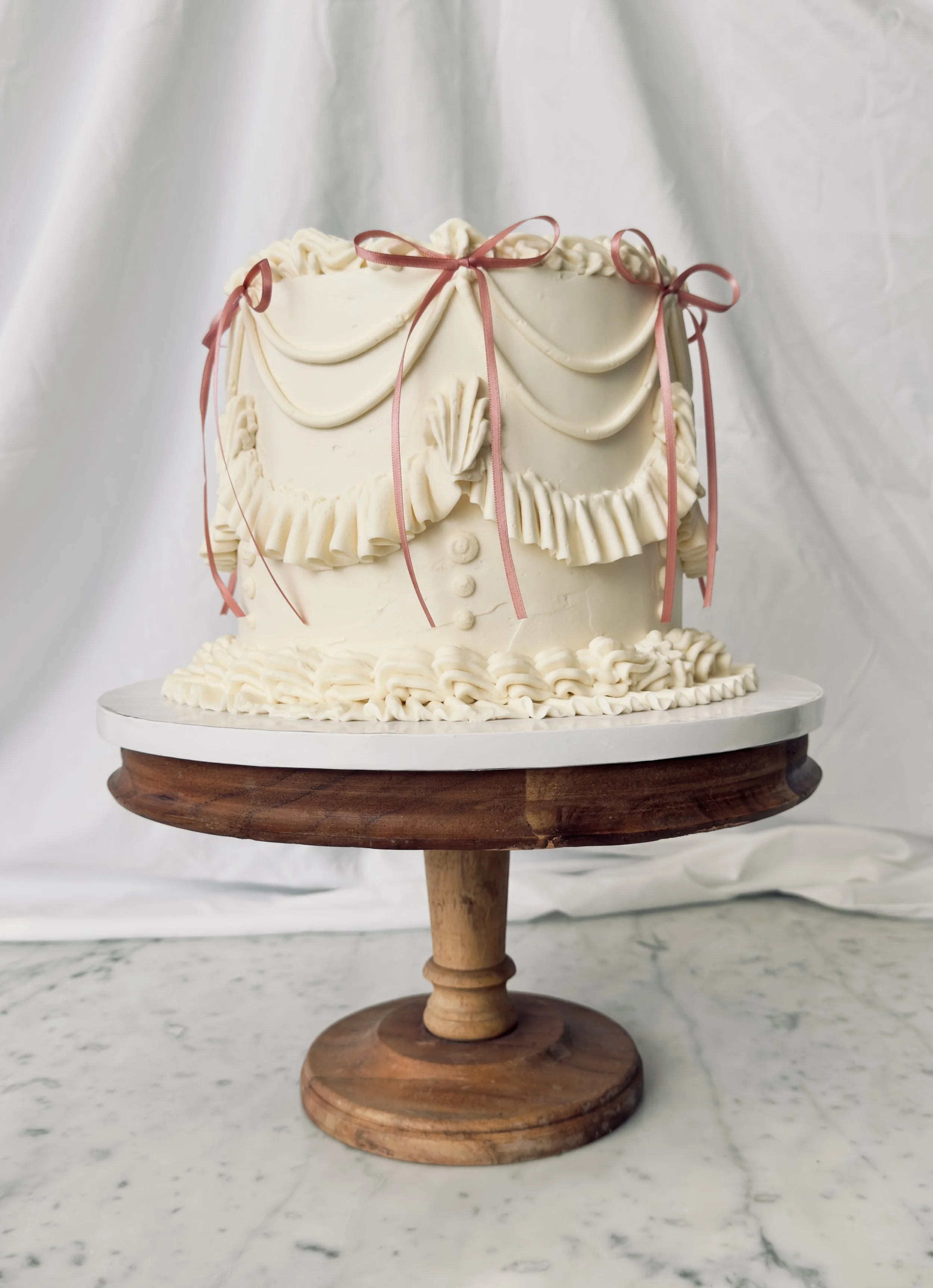 A round white cake decorated with ruffles, bows, and draped fondant, placed on a wooden stand against a white background.
