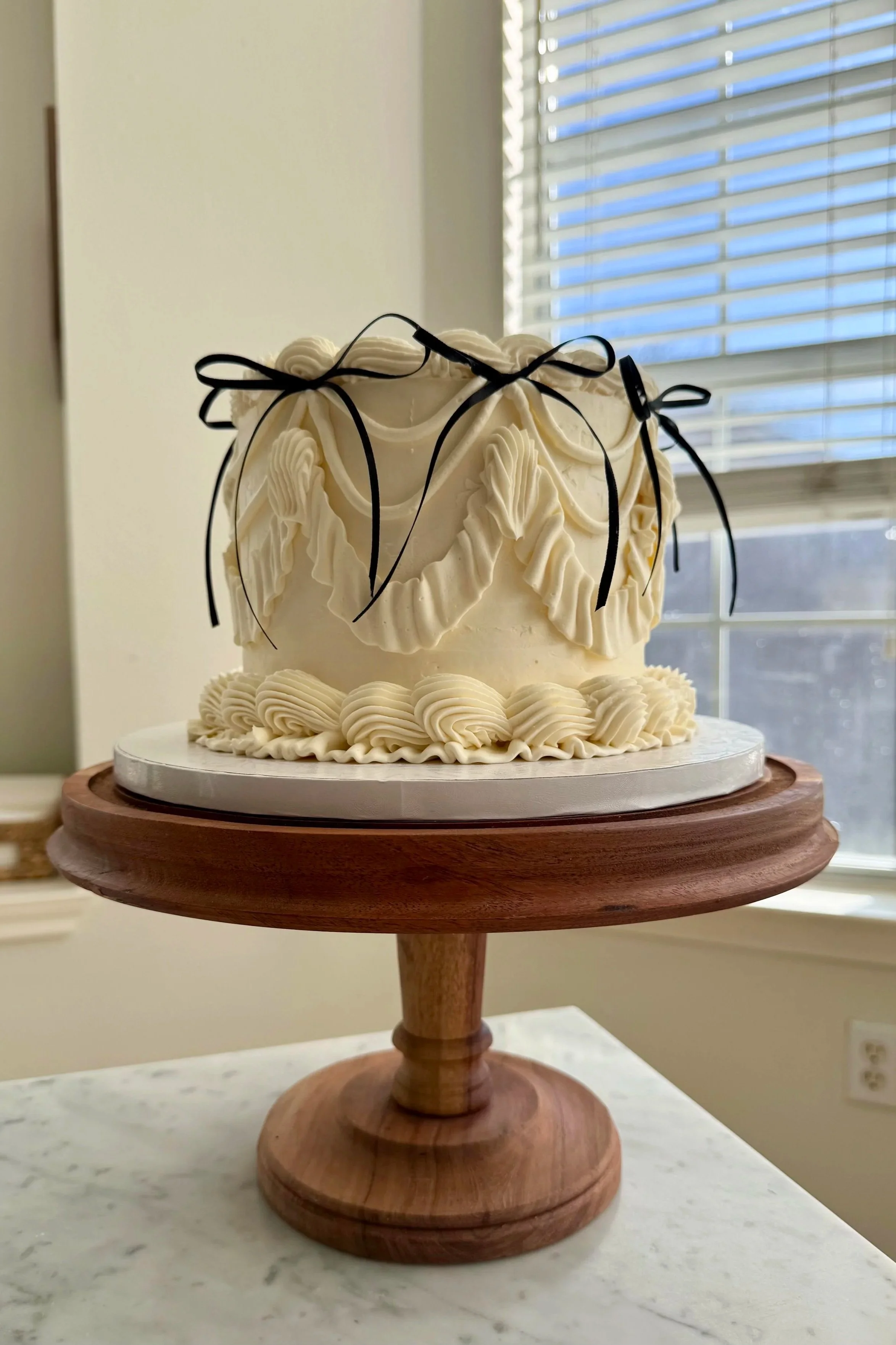 A white frosted cake with decorative piping, tied with black ribbons, on a wooden cake stand near a window with blinds.