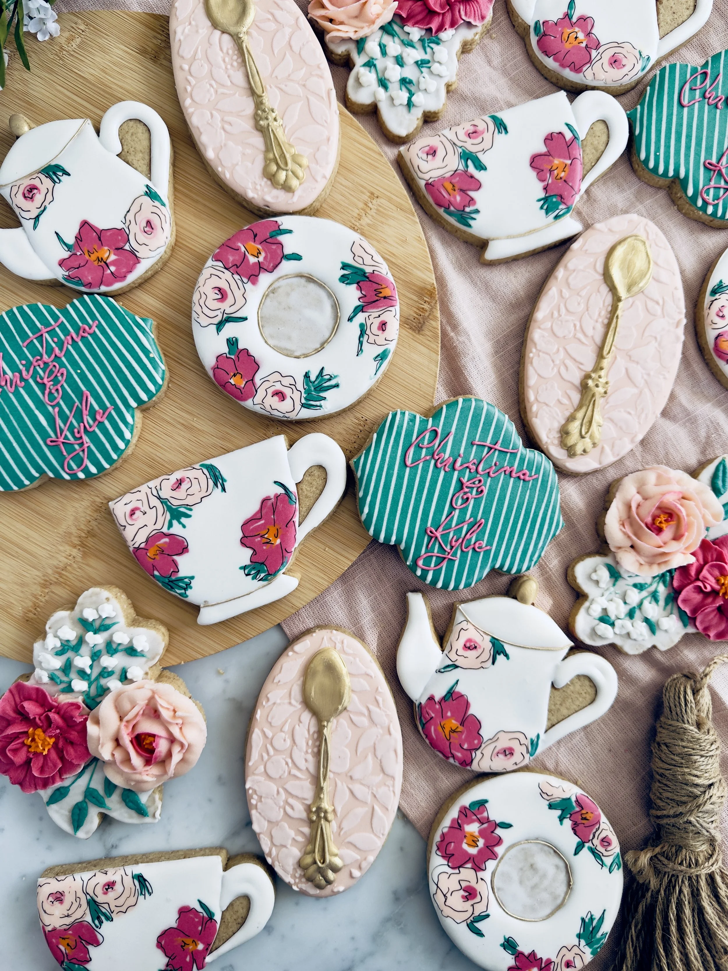 Decorative tea set cookies, including teapot, cup, plate, and spoon shaped cookies, decorated with floral and striped patterns, along with personalized cookies with names 'Christina & Kyle' and floral designs, arranged on a table with a pink cloth an