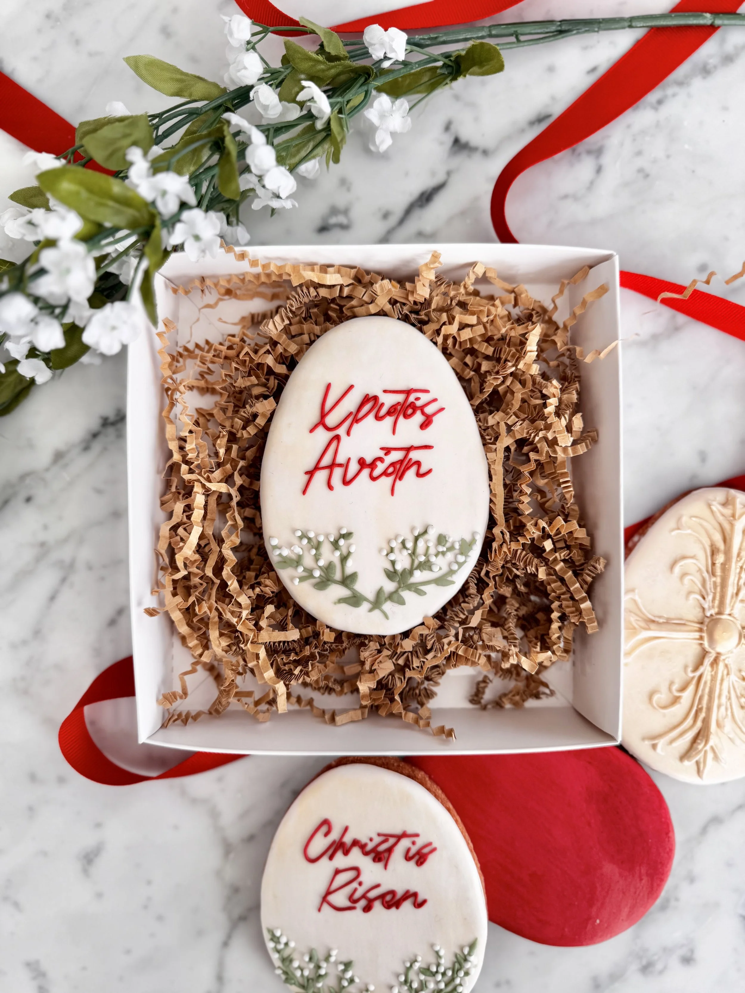 A white oval cookie with red writing and green floral decoration, placed inside a gift box with brown shredded paper, next to another decorated cookie and a red velvet heart.