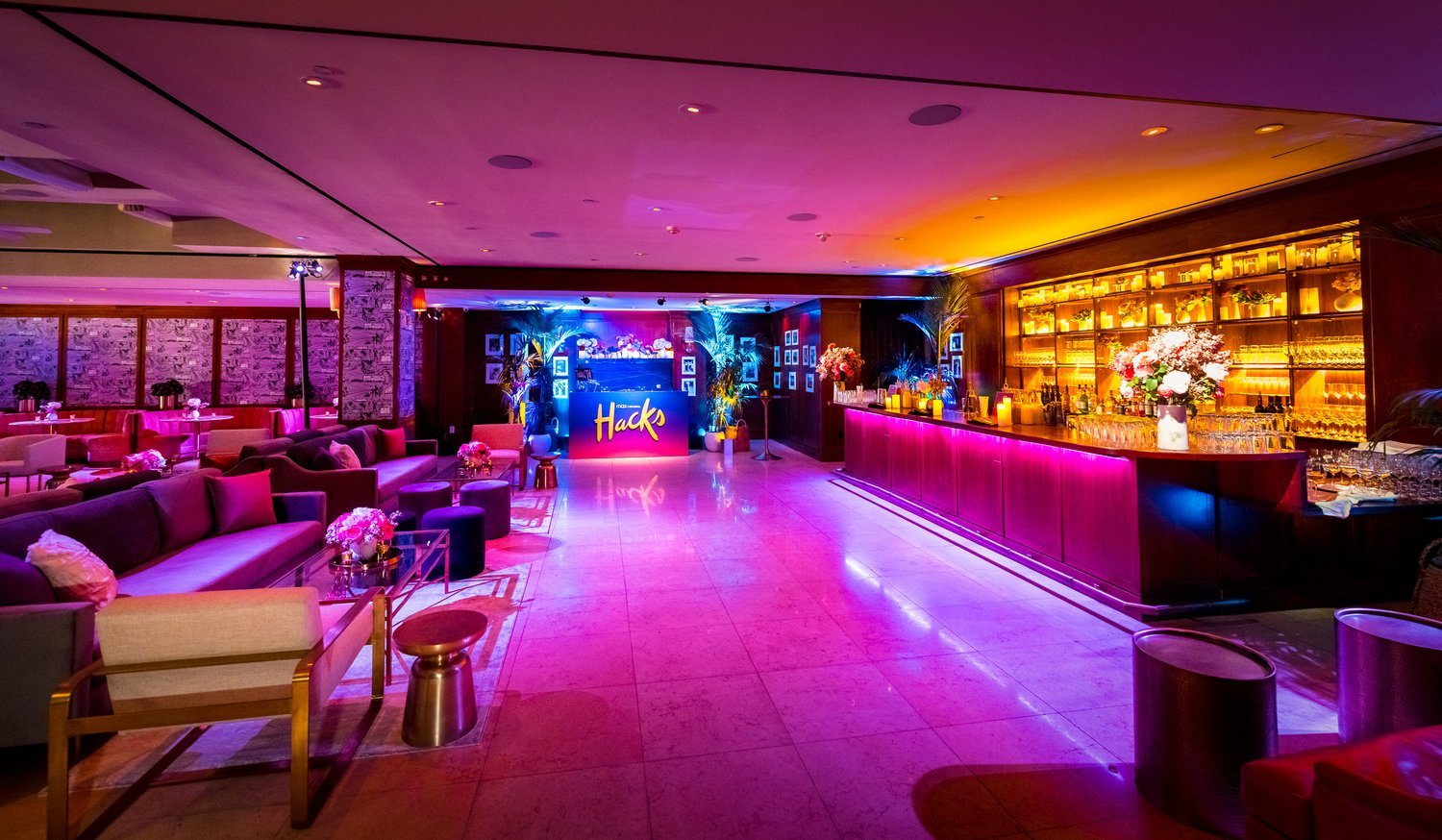 Luxurious hotel lounge with vibrant lighting, featuring comfortable seating, a purple-lit bar with glasses and bottles, and a floral arrangement.