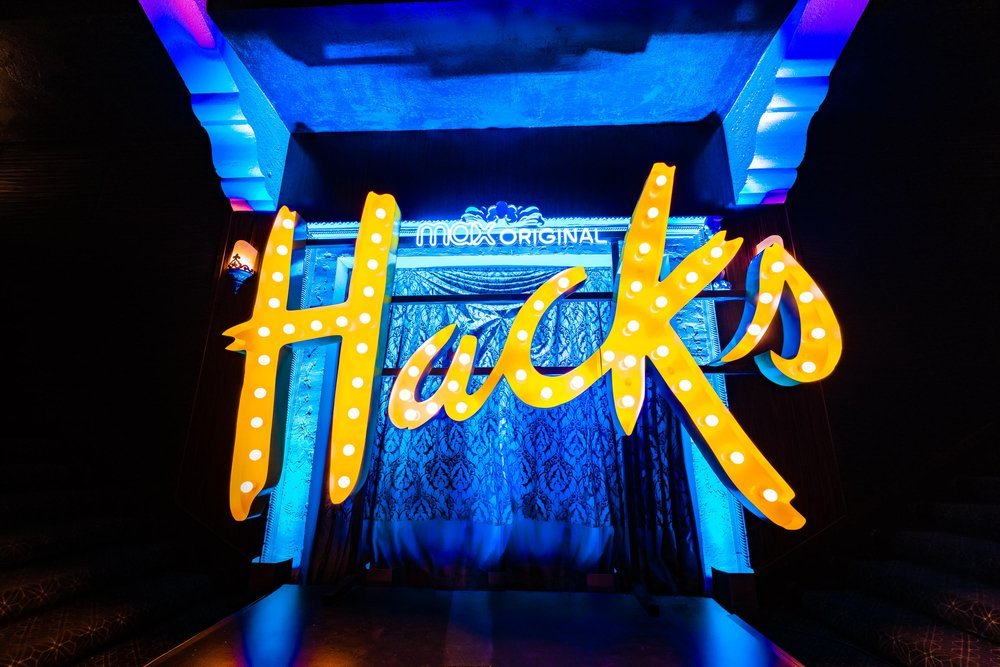 Colorful marquee sign with the word 'Hacks,' featuring illuminated yellow letters with white bulbs, set against a blue-lit background.