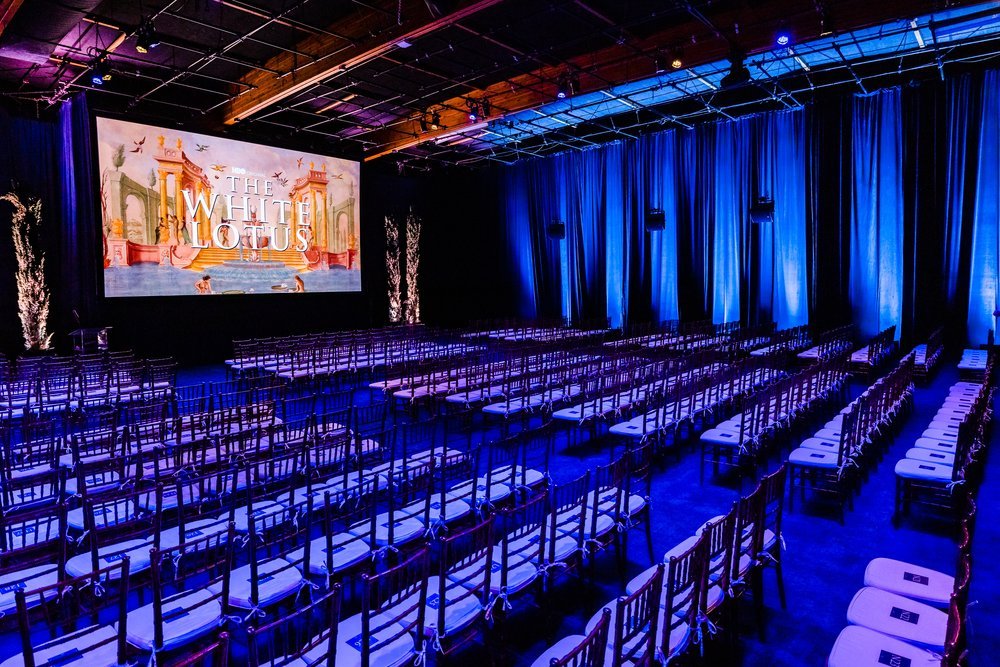 Event venue with rows of chairs, large screen displaying 'The White Lotus', blue lighting