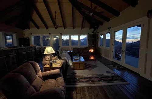 Cozy Hilltop House living room at Baxter Ranch featuring exposed wood beams, comfortable seating, warm lighting, and sunset views through large windows overlooking the mountain landscape.