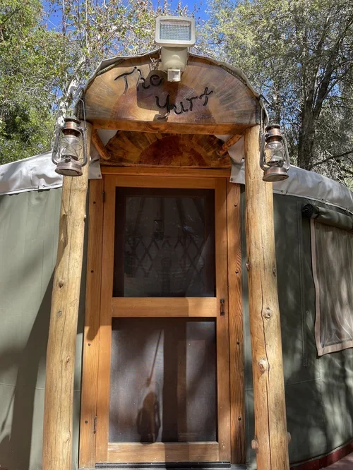 Entrance to The Yurt at Baxter Ranch, featuring a rustic wooden frame, screen door, and peaceful forest setting surrounded by trees and mountain scenery.