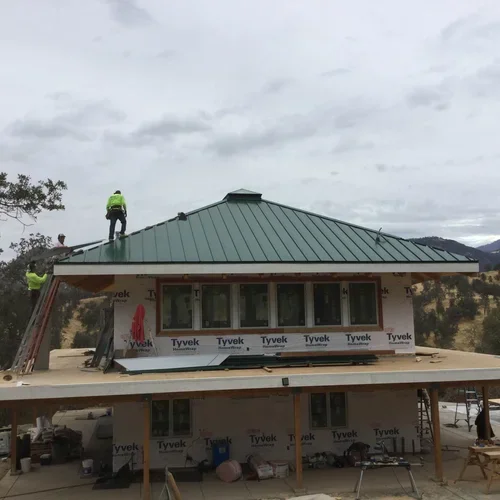 The construction of Hilltop House at Baxter Ranch, as builders install the green metal roof that would become part of the handcrafted mountain retreat surrounded by forest.