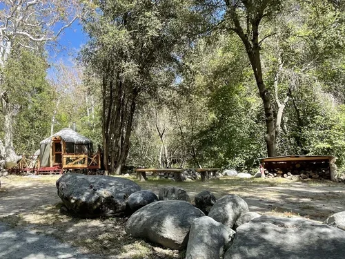 Outdoor camping area at Baxter Ranch featuring a teepee tent, picnic tables, natural rock features, and tall trees beneath the mountain sky.