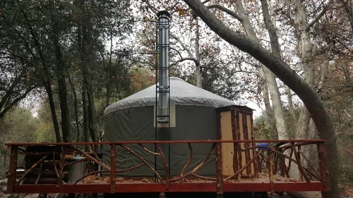 Exterior view of the Creekside Yurt at Baxter Ranch, featuring a private wooden deck, outdoor railing, wood-burning chimney, and peaceful forest surroundings.