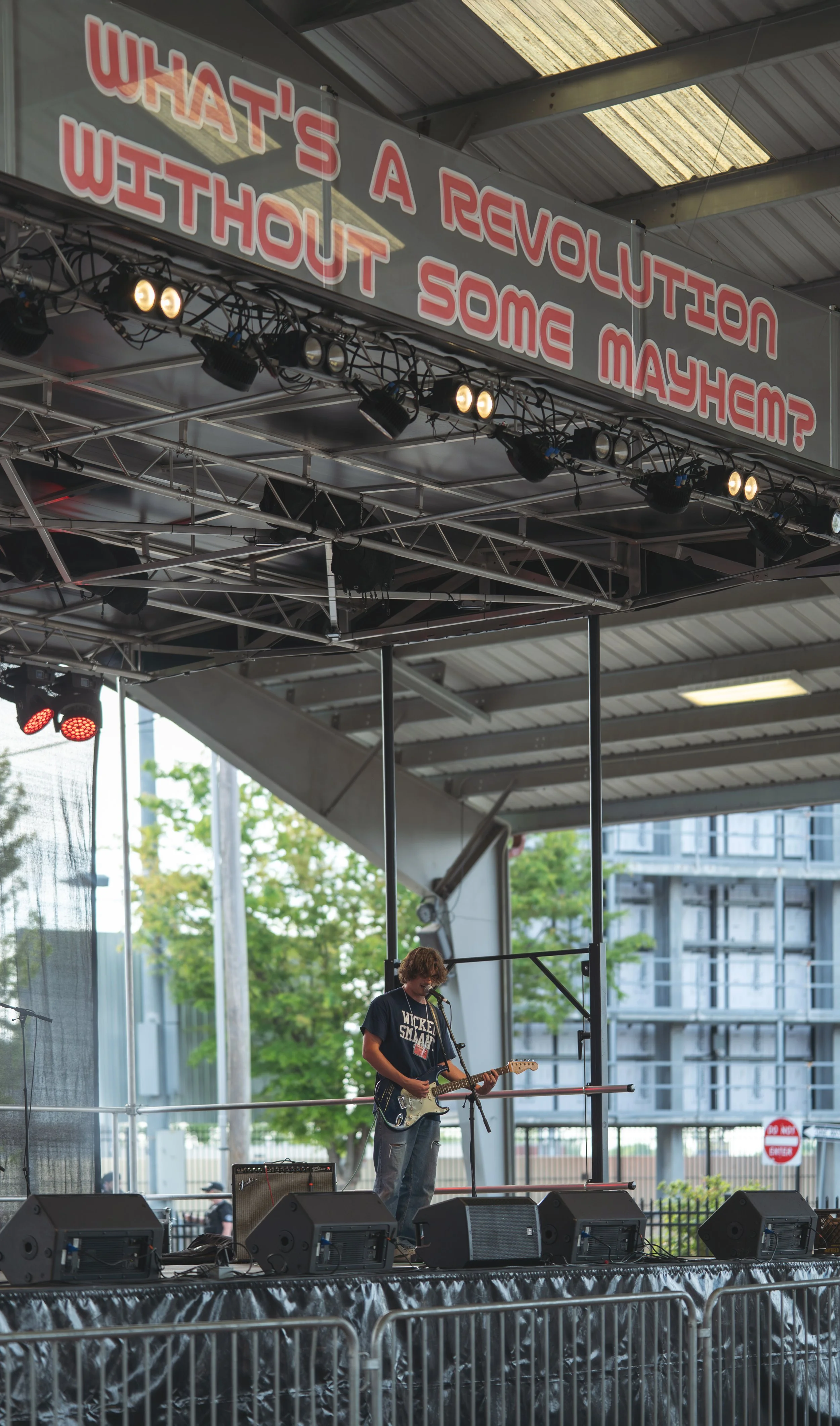 A person standing on an outdoor stage playing an electric guitar and singing into a microphone. There are speakers and stage lights, along with a large sign overhead that reads, 'WHAT'S A REVOLUTION WITHOUT SOME MAYhem?'